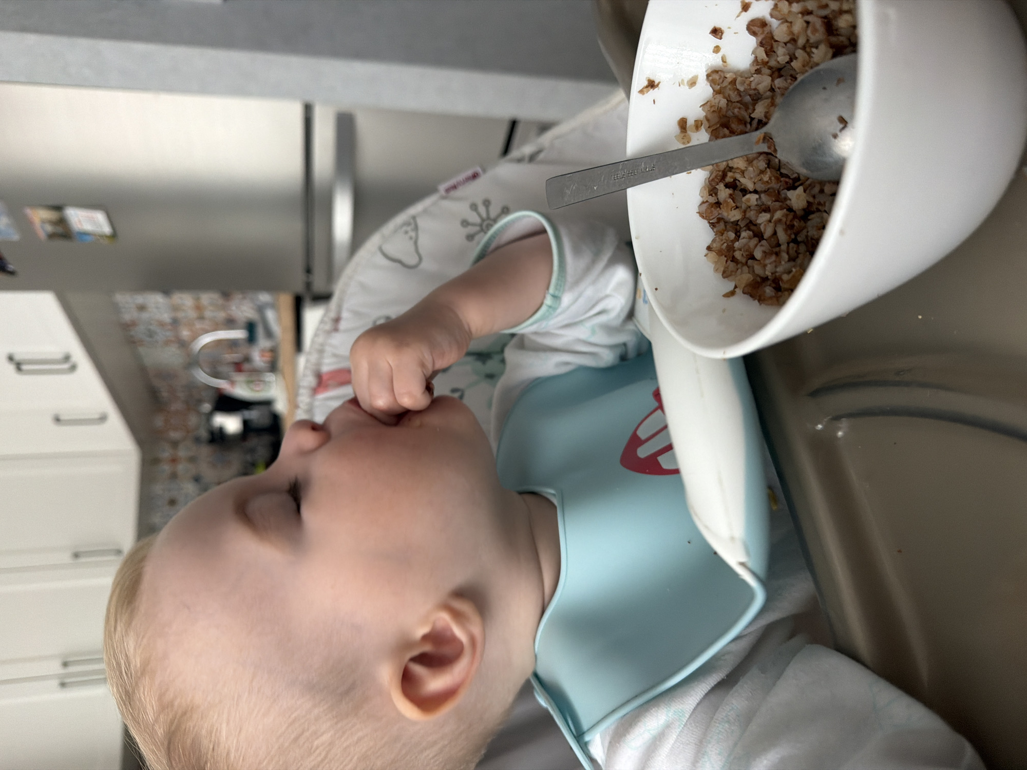 A baby with blond hair and a blue bib is sitting in a high chair and eating from a white bowl using a silver spoon. The bowl contains cooked buckwheat. The baby is holding their fingers to their mouth. The location appears to be a kitchen, with white cabinets and a stainless steel refrigerator visible in the background. The kitchen's backsplash is a multi-colored tiled design. There is no definitive indication of being in Votkinsk, Russia, as no location-specific landmarks or details are visible.