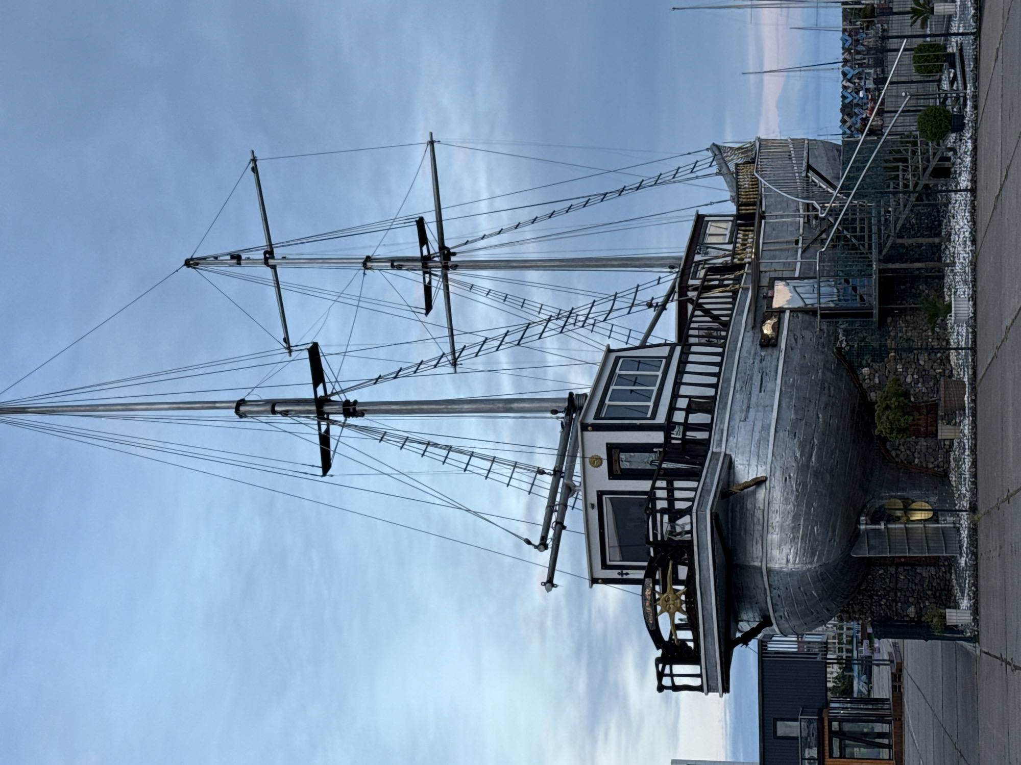 The image depicts a stationary ship replica, likely a building or structure designed to resemble a historic sailing vessel, positioned along a harbor or waterfront setting in Sirius, Russia. The ship features tall masts with rigging extending towards a cloudy sky. The hull is metallic in appearance, with visible window and cabin structures. A decorative ship's wheel is prominent. The ship's stern area is integrated with a building foundation. The surrounding environment includes other buildings, possibly a marina or port complex, and a paved surface.