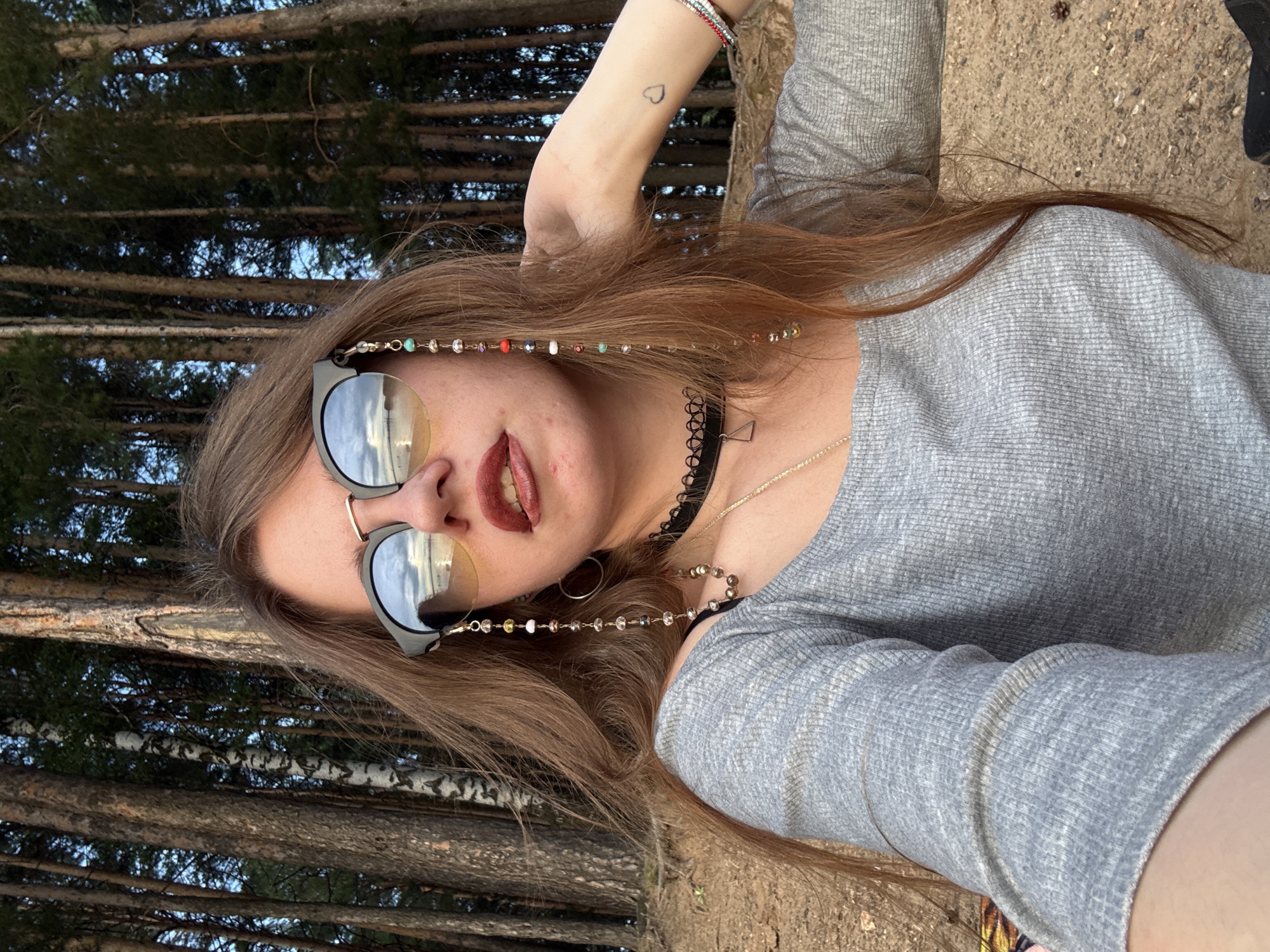 A woman with long, light brown hair is shown from the chest up, outdoors, with a forest of tall trees in the background. She is wearing sunglasses with a beaded chain attached to one lens. Her lips are painted a deep red, and she is wearing a grey long-sleeved shirt. She has a black choker necklace with a small geometric pendant. A small heart tattoo is visible on her left forearm. The ground in front of her appears to be sandy or dirt, with scattered pine cones. The lighting suggests it may be late afternoon or early evening.