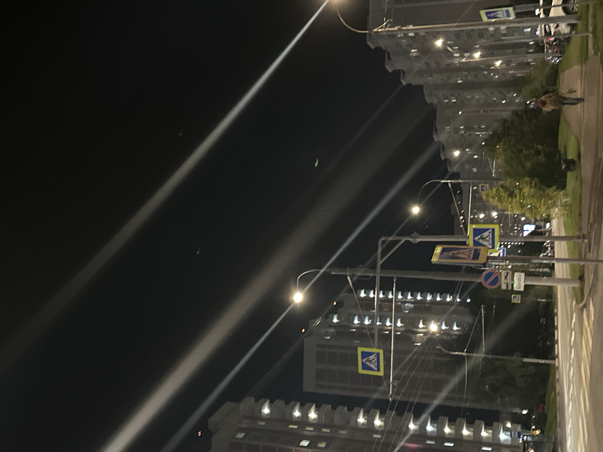 The image captures a nighttime scene in the city of Podolsk, Russia, likely on a street with apartment buildings and pedestrian crossings. Streaks of light, possibly from streetlights or illuminated signs, cut diagonally across the dark sky, creating a dramatic effect. In the foreground, a paved road with pedestrian crossings is visible, indicating a street or intersection. Several multi-story apartment buildings with illuminated windows line the street, suggesting a residential area. Various street signs, including a pedestrian crossing sign with a blue and yellow symbol and a red circular sign, are mounted on poles. Trees and green spaces are present along the sidewalk. A lone individual, walking away from the camera, is seen on the sidewalk. The overall impression is one of an urban environment at night, with artificial light sources creating distinct visual patterns against the dark sky.