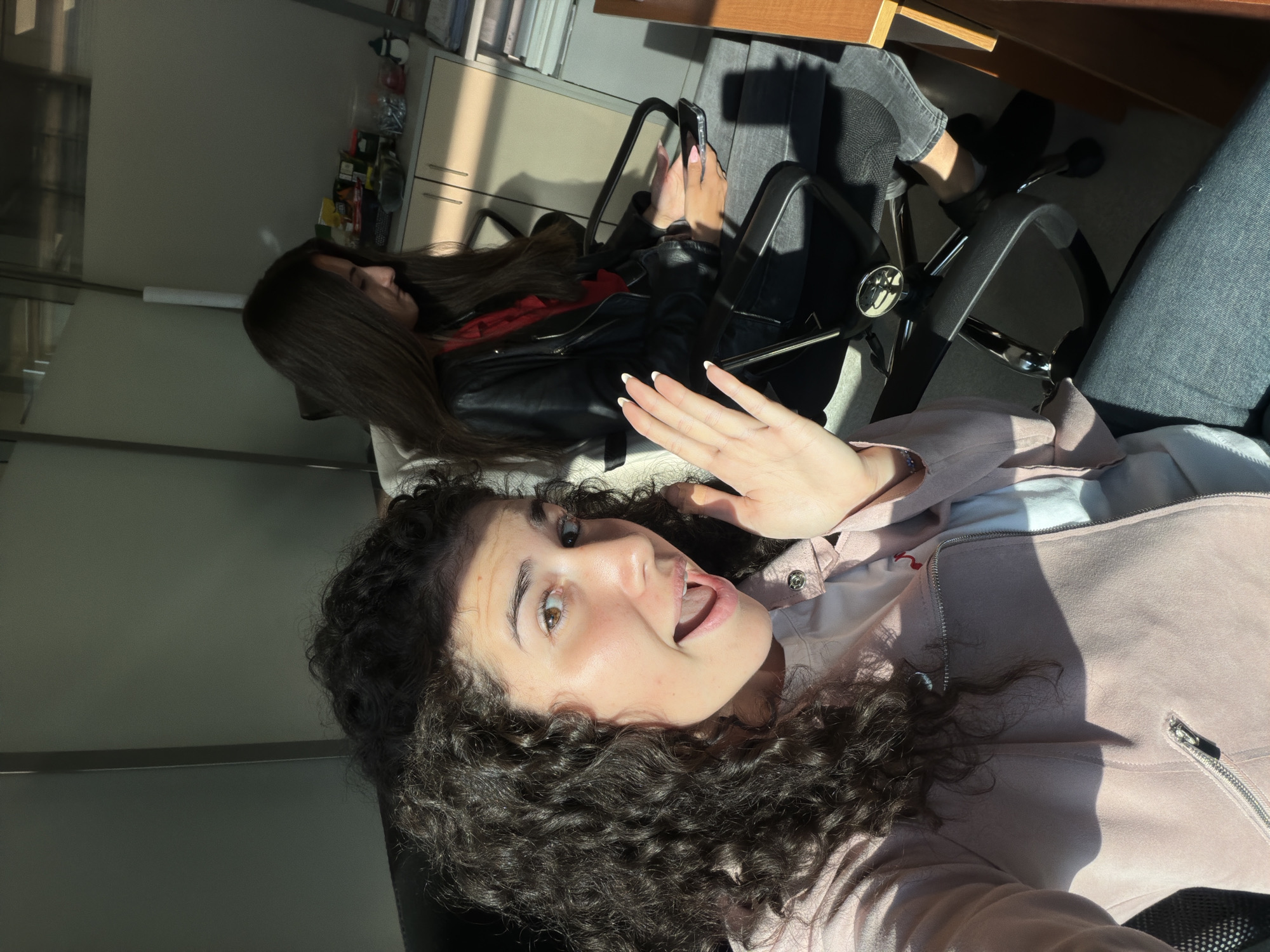 Two young women are seated in an office environment, illuminated by sunlight streaming from the left. The woman in the foreground, with dark curly hair, is facing the camera with her mouth open and her right hand raised, palm outward, in a gesture that could be surprise or greeting. She is wearing a light pink jacket over a white t-shirt and dark pants. The woman in the background, with long dark hair, is seated in a black office chair and looking down at her phone, which she holds in her left hand. She is wearing a black leather jacket over a red top and dark pants. Behind her, on a light-colored cabinet, are various items, including what appear to be snack wrappers, a clear container with small objects, and a small snowman figurine. The overall impression is a casual moment captured in an office setting. The scene is indoors, likely in an office building within the city of Sofia, Bulgaria, given the implied context of the request.