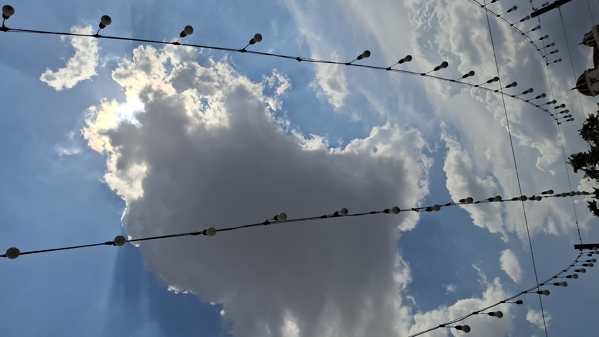 The image depicts a view of the sky with scattered clouds and the sun partially obscured by a large cloud formation. Several strings of outdoor lights, each with multiple incandescent bulbs, are strung across the frame, silhouetted against the sky. The lights are arranged in diagonal lines, creating a sense of depth. In the lower portion of the image, the tops of some trees and architectural elements of buildings are visible, suggesting an urban setting. Based on the metadata associated with the image, the location is identified as Shymkent, Kazakhstan. There are no people or specific actions clearly visible in the frame. The environment appears to be outdoors, likely in a public space or along a street, given the arrangement of the lights and the visible buildings.
