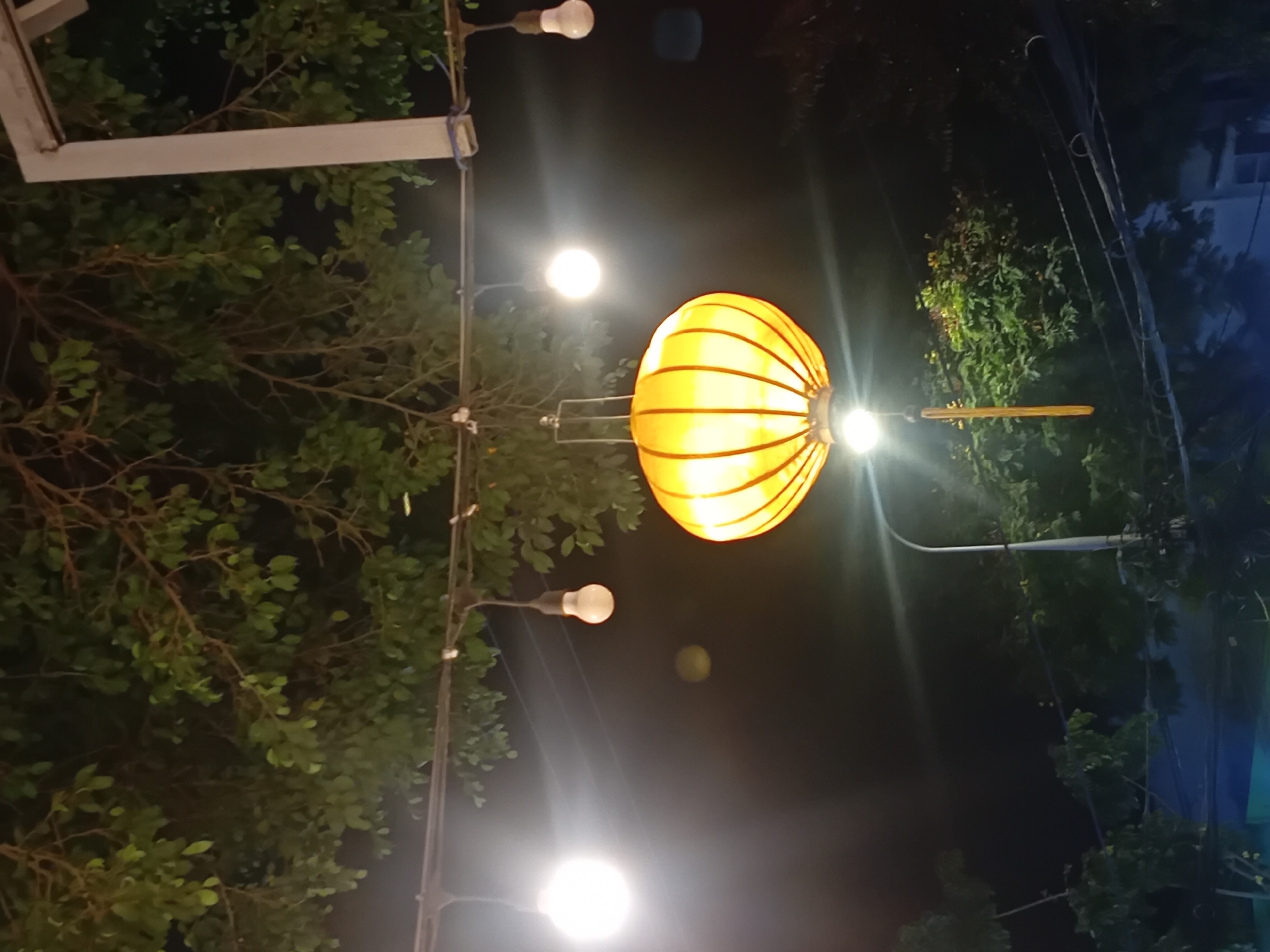 This image depicts an outdoor scene at night in Da Nang, Vietnam, possibly a street or an outdoor dining area. The focus is on a vibrant yellow lantern hanging from a string of lights, which are strung across the frame. Several other bare light bulbs are also visible, some glowing brightly and creating lens flares. Lush green foliage from trees overhangs the scene, providing a natural element against the artificial lighting. The background is dark, suggesting it's nighttime, with hints of distant structures and possibly other lights. The overall atmosphere is festive and warm, likely indicating a public space with decorative lighting.