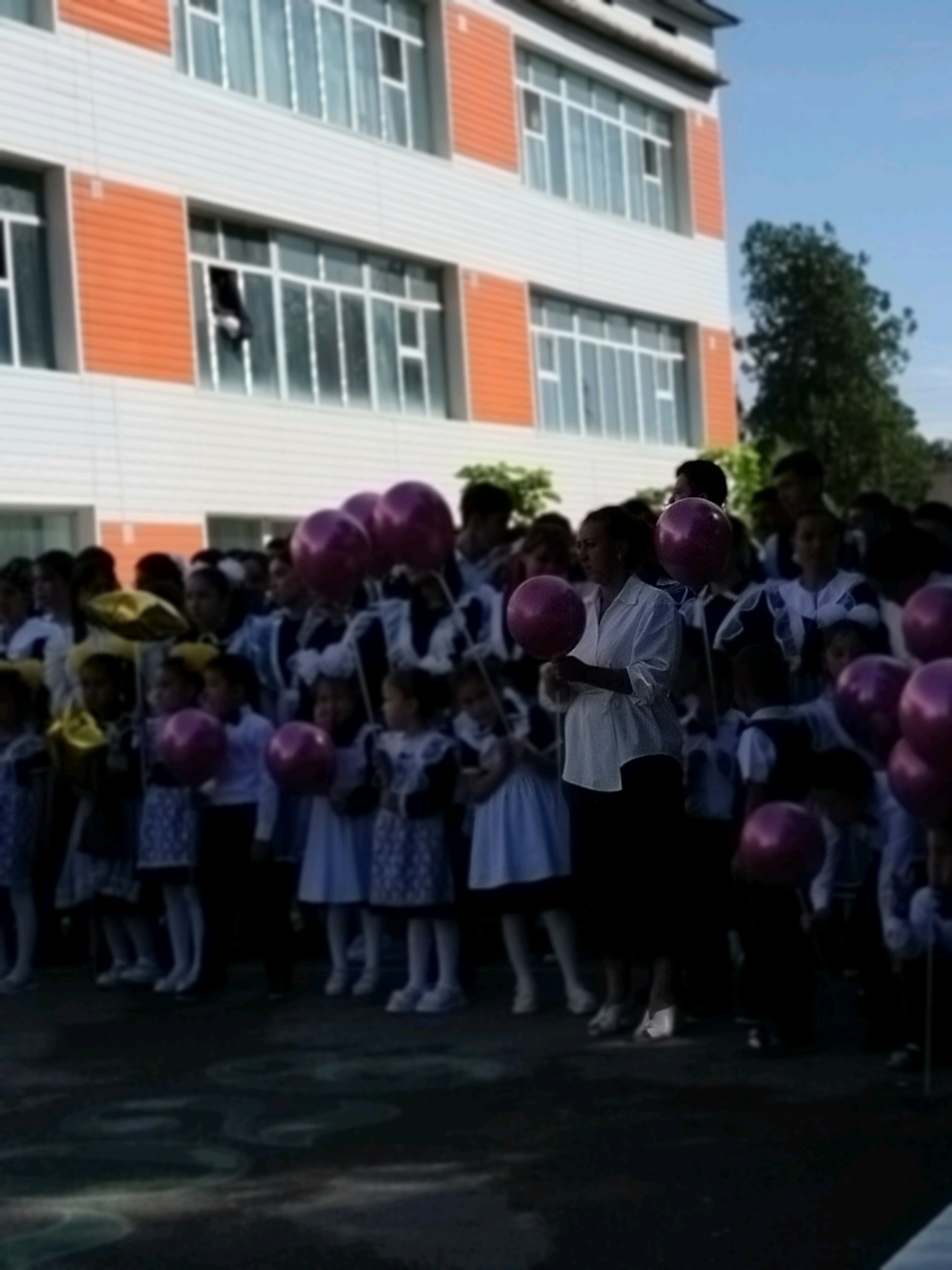 In the city of Kölkent, Kazakhstan, a group of children, presumably students, are gathered outdoors in front of a modern, multi-story building with orange and white facade elements and large windows. Many of the children are dressed in uniforms, some appearing to be school uniforms with blue and white elements. They are holding and interacting with numerous large, round, pink balloons. One adult woman, wearing a white blouse and dark skirt, is also present and holding a pink balloon. The ground in front of the building appears to be a paved area or courtyard, with some patterned markings visible, possibly chalk drawings or a decorative surface. The sky is clear and blue, indicating daylight. The overall scene suggests an event or celebration, likely related to a school.