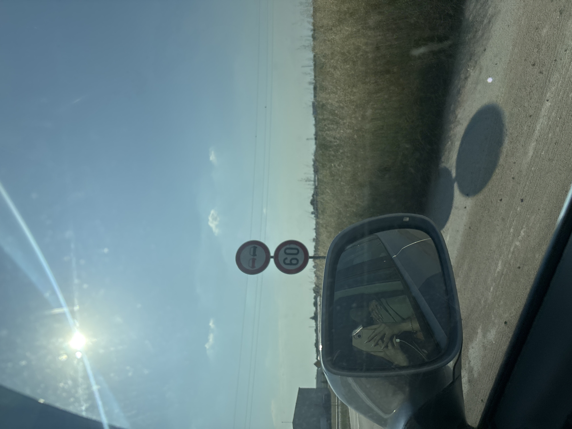 An outdoor image, captured from the interior of a vehicle, displays a bright sunny day with a clear blue sky. A strong sun flare emanates from the top-left corner of the sky, casting bright streaks across the upper portion of the frame. In the midground, two European regulatory road signs are stacked vertically on a pole. The top sign, a red circle with a white interior, features two black car silhouettes (one red, one black) with a diagonal line through them, indicating a "no overtaking" restriction. Beneath it, a similar red circular sign with a white interior displays the number "60" in black, signifying a "60 km/h" speed limit.
The landscape to the right consists of a vast field of tall, dry, golden-brown grass or crops, stretching towards a flat horizon. Power lines traverse the sky above this field. To the left, a large, dark-colored industrial building or warehouse is visible in the distance. The foreground on the bottom right shows a dirt or gravel path with two distinct, dark, circular shadows and a small bright white light reflection.
The vehicle's passenger-side mirror occupies the lower-left quadrant of the image. Reflected in the mirror is the driver or passenger holding a light-colored smartphone (equipped with multiple rear cameras) to capture the photograph. The person's hand, adorned with long, light-colored nails, is prominent, holding the phone which has a black charging cable plugged into its port. A portion of the person's upper body, wearing a light-colored garment, is also reflected, along with details of the car's interior. The scene is located in Stryama, Bulgaria.