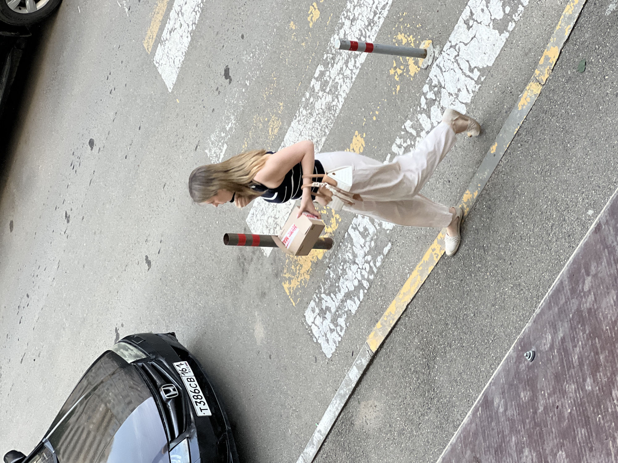 A woman is walking across a crosswalk on a paved city street. She is wearing a black and white striped sleeveless top, light-colored wide-leg pants, and light-colored flat shoes. She is carrying a small brown box in her right hand and a small white handbag with tan accents in her left hand. A black car with a license plate reading "T386CB 161 RUS" is parked on the left side of the image. Red and white striped bollards are present near the crosswalk. The ground is asphalt with white and yellow painted lines indicating the crosswalk. The image appears to be taken from an elevated perspective.