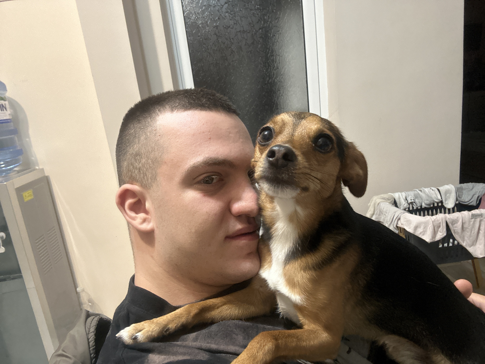 A young man with a buzz cut is holding a small brown and black dog in his arms. The dog is looking directly at the camera with its front paws resting on the man's chest. The man is looking at the camera with a neutral expression. In the background, there is a window, a water cooler, a cabinet, and a laundry basket filled with clothes. There is no clear indication that the photo was taken in Burgas, Bulgaria.
