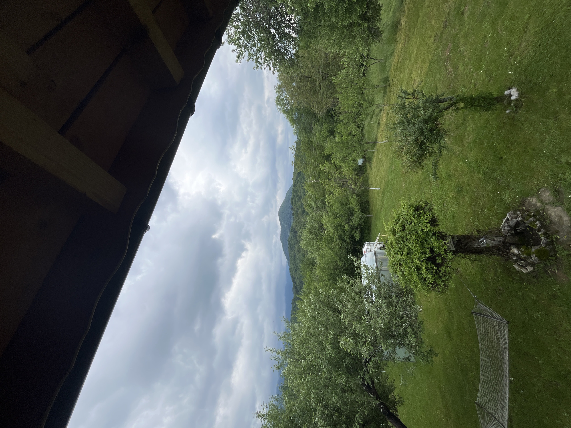 The image shows a view from a structure, possibly a cabin or porch, looking out over a green landscape in Shopite, Bulgaria. The left side of the frame is dominated by the wooden structure's roof. The view opens onto a cloudy sky above a mountain range. Below, a grassy field is punctuated by trees and a small building. A hammock is visible in the lower right.