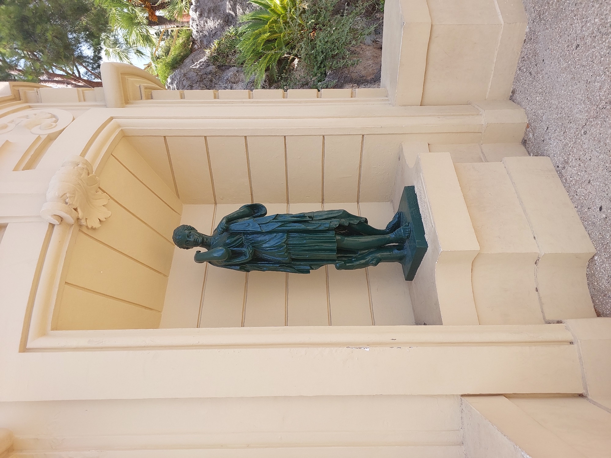 This is a full shot photograph of a dark green bronze statue of a draped female figure, likely ancient Greek or Roman, standing in a recessed alcove. The alcove is part of a pale yellow ornate building with classical architectural elements, including decorative carvings above the alcove. The statue is positioned on a raised, multi-tiered pedestal within the alcove. Behind the alcove, to the right, is a rocky incline with lush green vegetation, including palm trees and other plants, indicating an outdoor setting. The ground in front of the alcove is covered with small, light-colored pebbles. The overall impression is a garden or public space within a city. The architecture and landscaping suggest a location in a warm climate, consistent with Monaco.