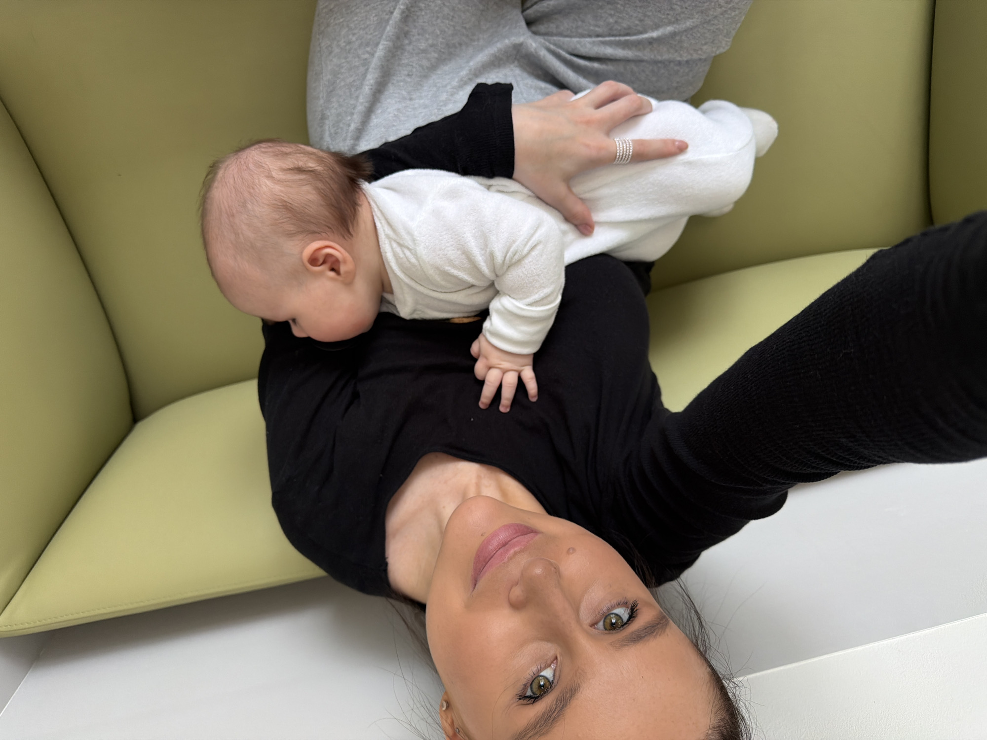 A young woman is lying on a light-yellow couch while holding a baby on her chest. The woman is wearing a black long-sleeved shirt and gray sweatpants. She is smiling at the camera. The baby is wearing a white onesie. The background is out of focus. The location cannot be determined from the image.