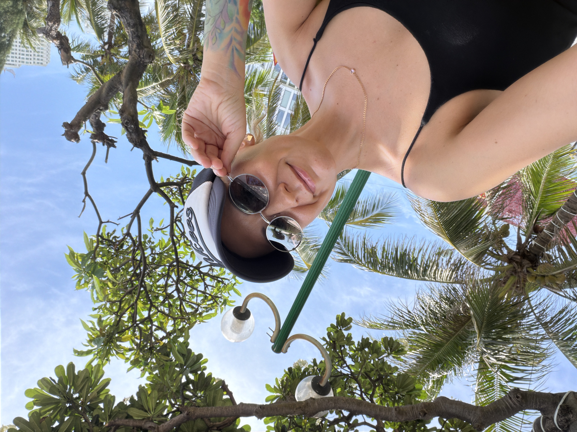 A woman wearing a black tank top, a black and white baseball cap, and sunglasses takes a selfie outdoors. She is looking up and to her right, adjusting her sunglasses with her right hand. She has a delicate gold necklace with a small pink pendant. Her left arm is bare, revealing a colorful tattoo of leaves on her forearm.
The background shows a bright blue sky with scattered clouds. There are lush green palm trees and other tropical foliage. A street lamp with two globes is visible above and behind her. In the distance, a tall, white building can be seen, indicating an urban setting within a tropical environment. The scene is consistent with a location in Nha Trang Ward, Vietnam, known for its beaches and vibrant city life.