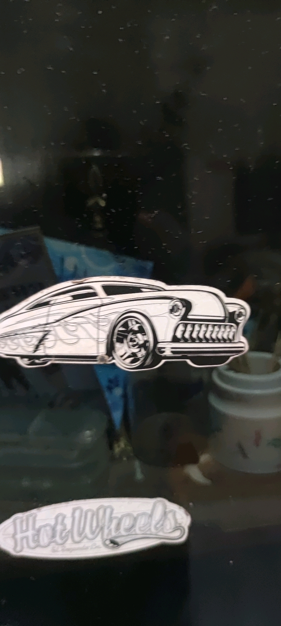 The image displays a sticker of a classic car, likely a Hot Wheels brand due to the "Hot Wheels" logo sticker also present. The car is depicted in a stylized black and white outline with flame-like details on the side. The background is dark and appears to be a reflective surface with small specks, possibly dust or water droplets. There is a hint of blue fabric or material behind the car sticker and a blurred image of a white ceramic object to the right. The sticker of the car is positioned in the center of the frame, with the "Hot Wheels" logo sticker below it and to the left. The image is taken in a way that suggests the stickers are affixed to a flat surface. There is no discernible information about the city of Gambarsari, Indonesia within the image itself.