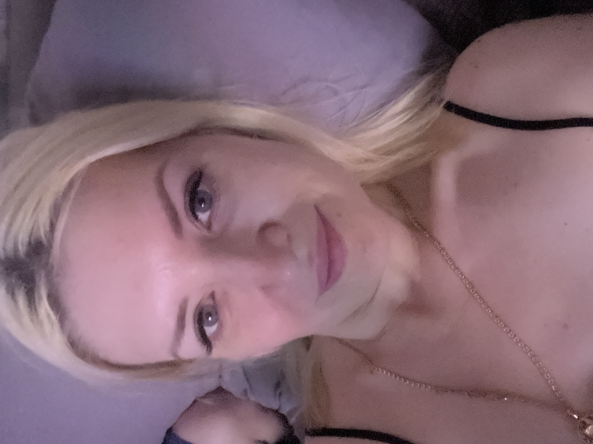 A close-up, overhead, selfie-style shot shows a blonde woman with light-colored eyes and pink lipstick lying down on a surface that appears to be a bed. She is wearing a black spaghetti strap top and a gold necklace. The background is out of focus and appears to be a pillow or blanket in shades of purple and pink. There is a slight tilt to the camera angle, and the lighting is dim, casting a pinkish hue over the scene. The location is identified as Tyumen, Russia, though no specific landmarks or details within the city are visible in the image itself.