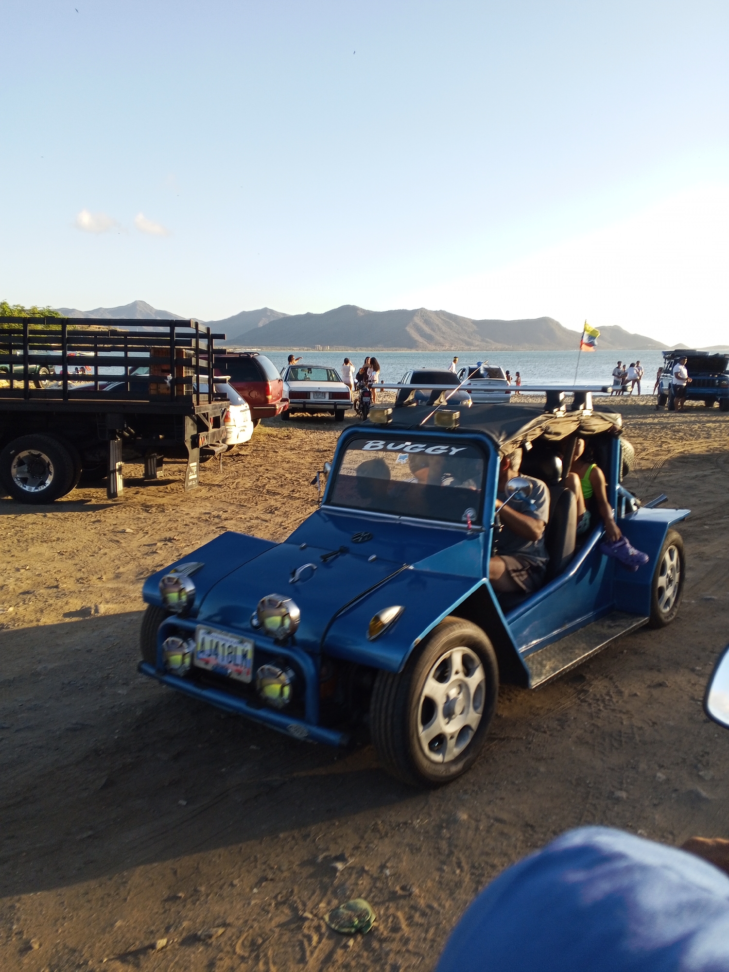 A blue buggy vehicle is prominently featured in the foreground, parked on a sandy beach. The buggy has the word "Buggy" written across the top of the frame with a Venezuelan license plate: 1A18LM. The buggy contains two people in it. In the background, there is an ocean with boats and other beachgoers. Various cars and trucks are parked in the beach area. The scene is captured under a clear sky with the sun positioned high, as the shadows suggest. The location is identified as the city of Juan Griego, Venezuela, based on the Venezuelan flag in the background.