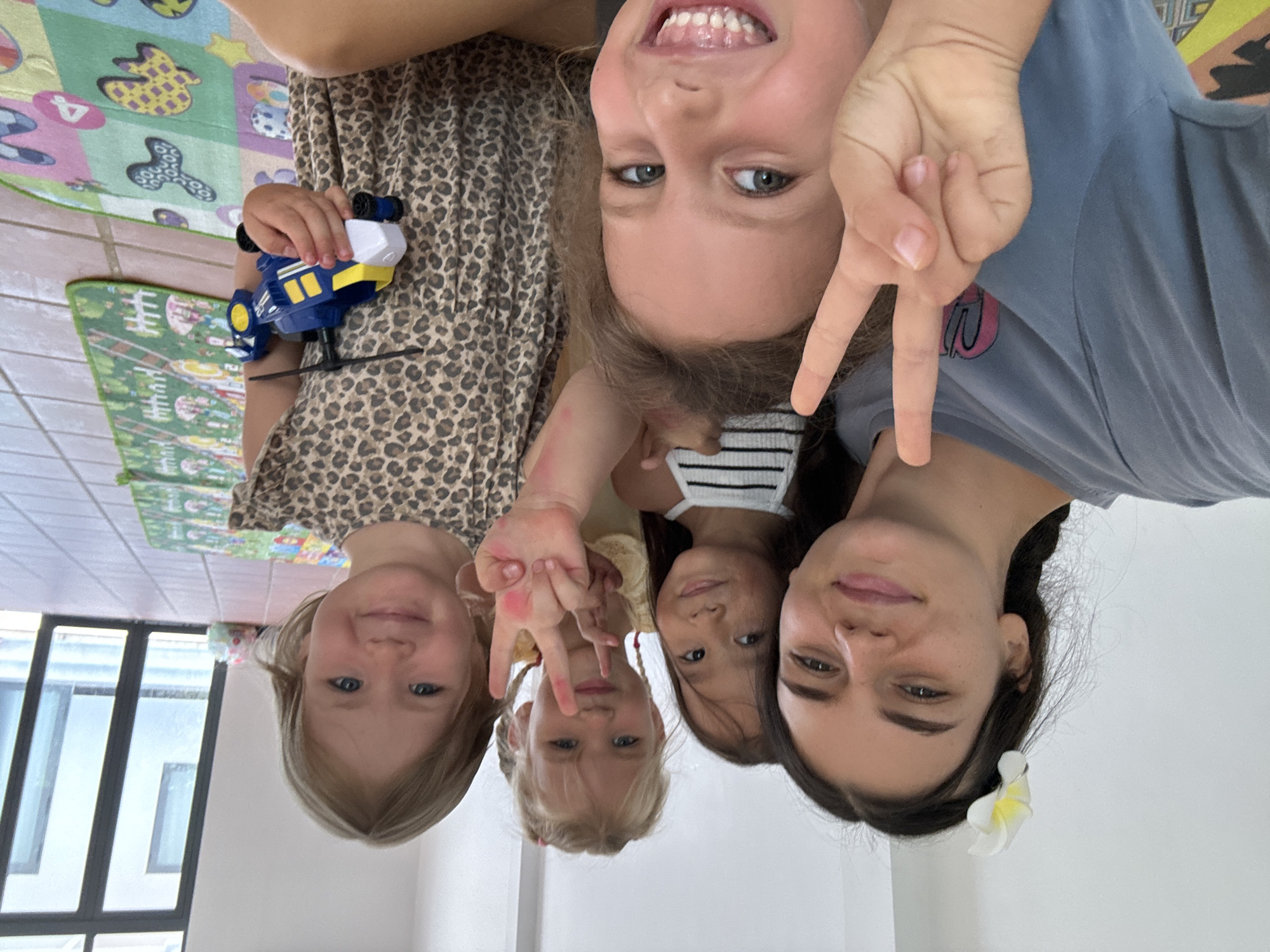 A photo shows four girls and a woman, apparently close together, with the camera looking up at them. The blonde-haired girl at left wears a leopard-print dress and holds a blue and yellow toy helicopter. Above her, another girl smiles broadly, her teeth visible. The girl in the center wears a striped top. The woman is to the right, wearing a blue top and has a white flower in her dark hair. Two girls are making peace signs with their fingers. The setting looks like an interior, possibly a house in Mũi Né, Vietnam, judging by the white walls and light. In the background, a colorful children's play mat is visible, and a window allows natural light into the room.