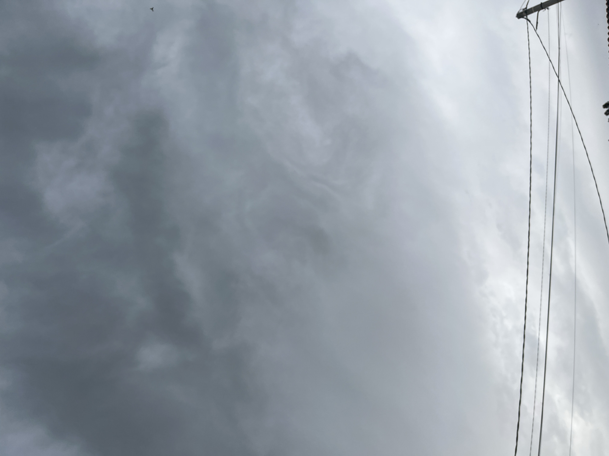 The image depicts a cloudy sky with a few power lines stretching across the lower portion. The clouds are dark and overcast, suggesting a potential for rain or a stormy day. In the far upper right corner, a small bird is visible in flight. The power lines are suspended between poles, some of which are visible on the right side of the frame. The overall scene is urban, indicated by the presence of the power lines and the implied infrastructure. The location of Romanovka, Kyrgyzstan, is not visually evident from the image itself, as there are no specific landmarks or identifying features to confirm this location.
