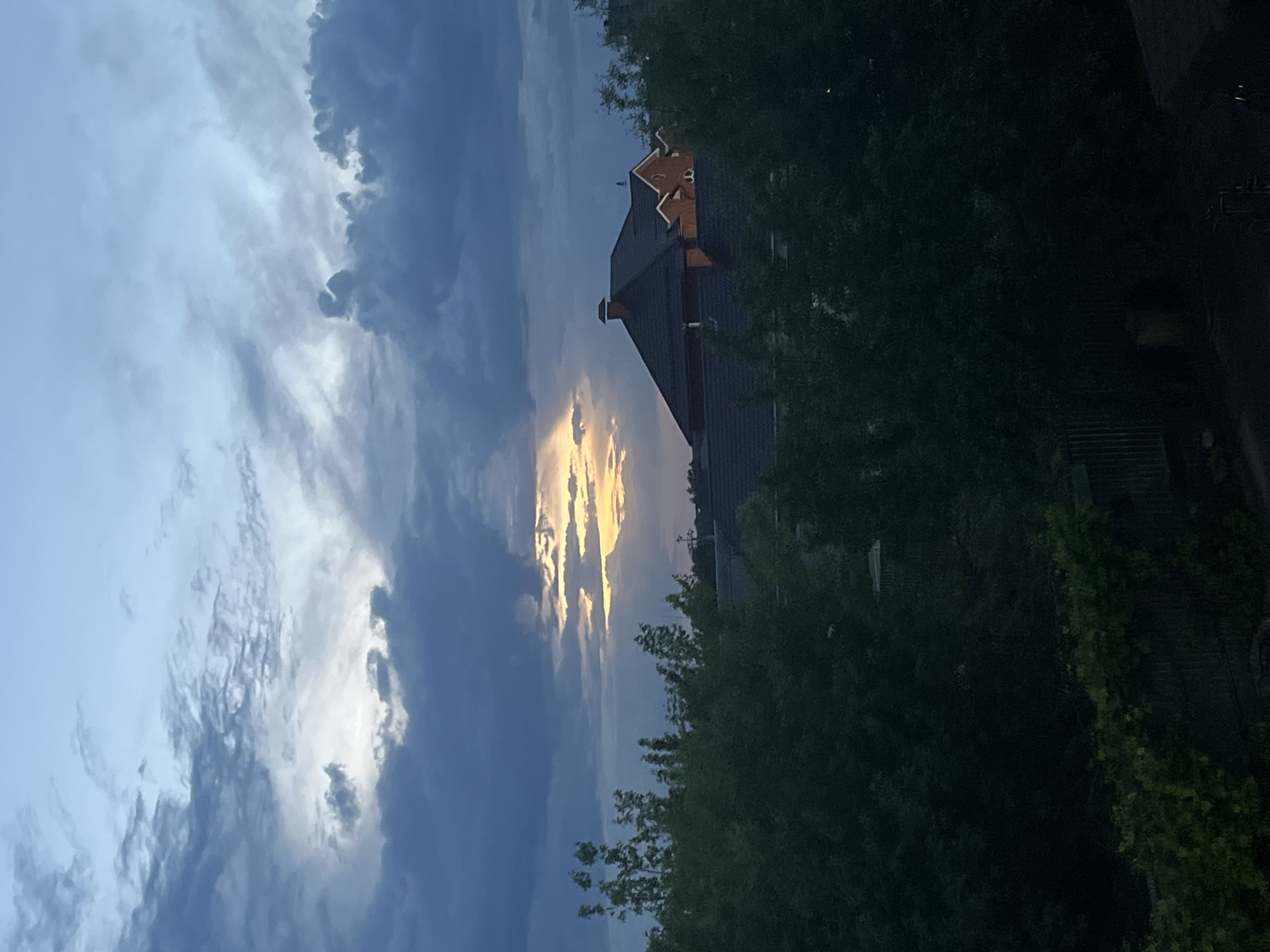 The image depicts a sky with dramatic clouds, a house, and trees. A bright, golden light shines through a gap in the clouds. The house is dark in color with a brown roof, with a gable decoration. The trees are dark green and partially obscure the house. The location is unknown. There are no people visible in the image.