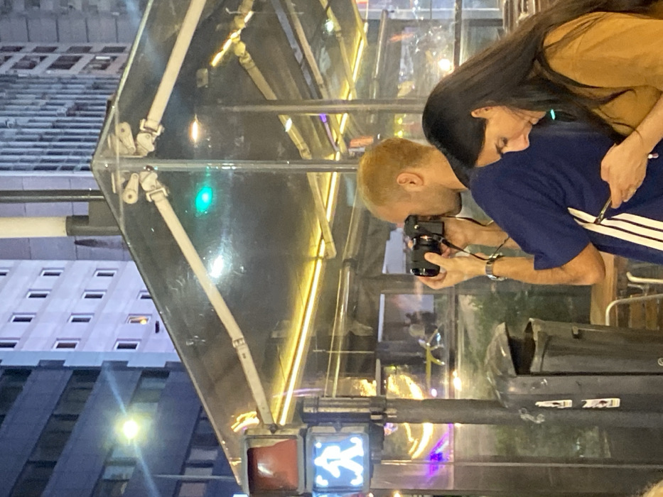 A man with blonde hair is taking a picture with a black camera. A woman with long dark hair is standing behind him, embracing him. They appear to be outdoors, possibly in a city setting. A clear structure with lights and metal supports is visible, with a pedestrian traffic light showing a walking symbol in the corner. Buildings are reflected in the clear structure, indicating the scene is likely at night.