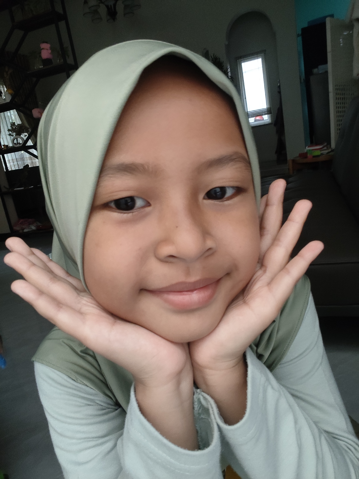 A young Indonesian girl, wearing a light green hijab and a long-sleeved shirt of the same color, is shown in a close-up portrait. She is resting her chin on her hands, which are placed against her cheeks, and she is looking directly at the camera with a gentle smile. Her eyes are dark and her skin tone is light brown. The background appears to be an indoor setting, possibly a living room, with shelves containing items and a window visible in the distance. The lighting is natural, casting soft shadows. The image is likely a selfie, taken by the girl herself. The location is unspecified, and there is no information to suggest it is in Pemalang, Indonesia.