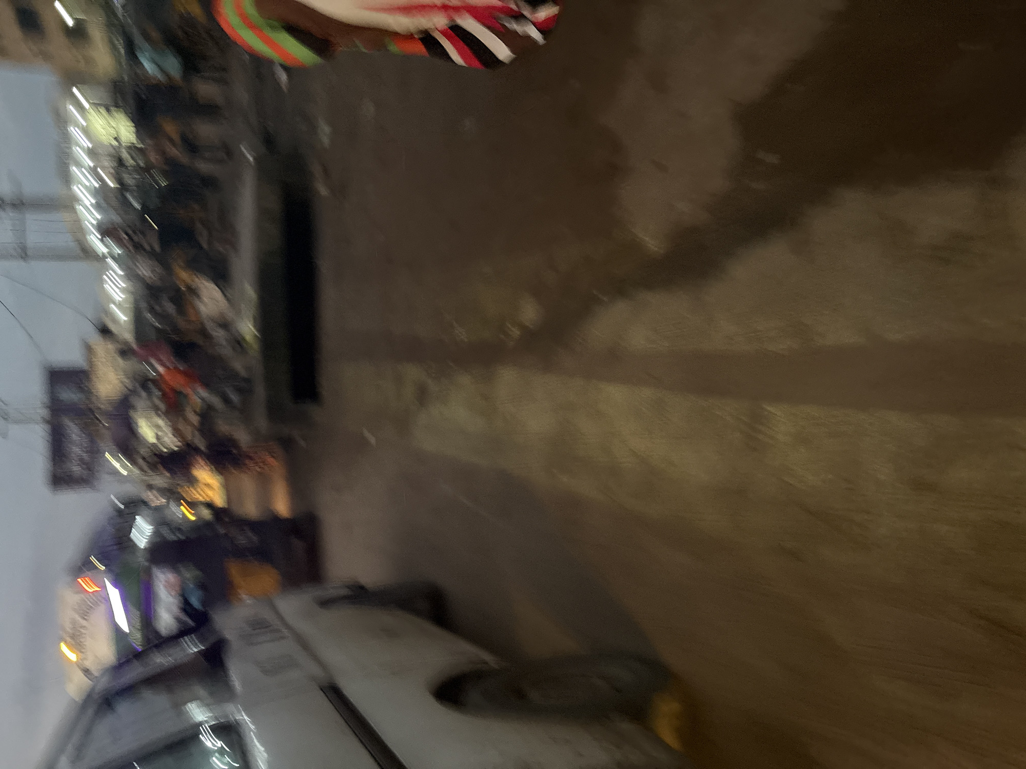 This image depicts a street scene in Onitsha, Nigeria, likely at dusk or in low light conditions. A white van is partially visible on the left side of the frame, facing away from the viewer. The van appears to be stationary or moving slowly. In the background, a busy street is evident with multiple people and vehicles. Lights from buildings and vehicles create streaks due to motion blur. Some individuals are seen standing or walking, and there appears to be a market or roadside stalls. The ground is a dirt or paved surface, showing signs of wear and tear. A person wearing colorful clothing is partially visible on the right edge of the image. The overall impression is one of a bustling urban environment in Nigeria.
