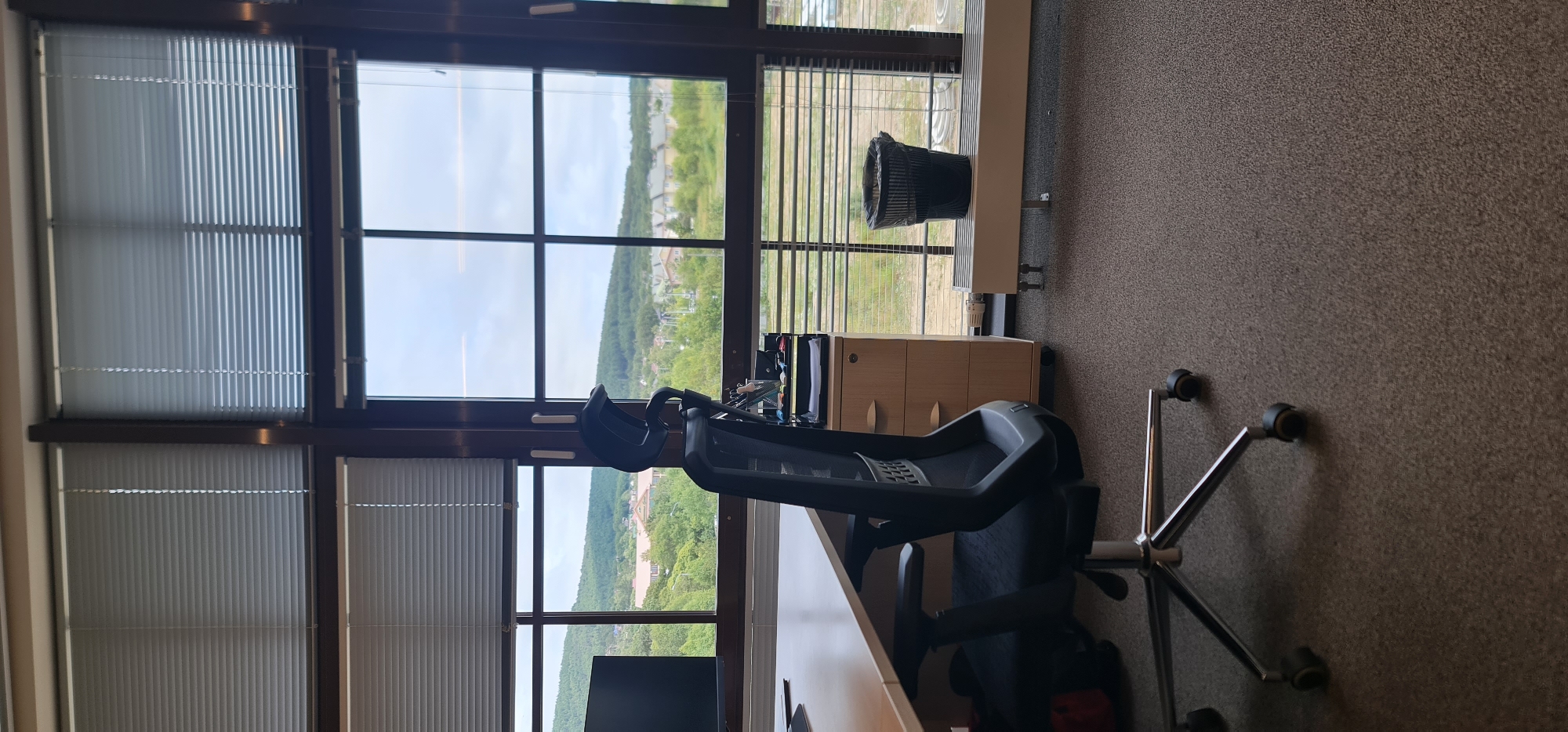The image depicts an interior view of an office with a large window overlooking a landscape. In the foreground, a black office chair with a metal base and casters is positioned in front of a light-colored desk. To the right of the desk, there is a rolling filing cabinet with three drawers. A black trash can with a liner sits on a long, white radiator beneath the window. The window is divided into four panes and covered with horizontal blinds that are partially open, revealing a view of rolling green hills, houses, and trees under a cloudy sky. The office is carpeted in a dark brown, textured material. The scene appears to be in Yuzhno-Sakhalinsk, Russia, based on contextual information.