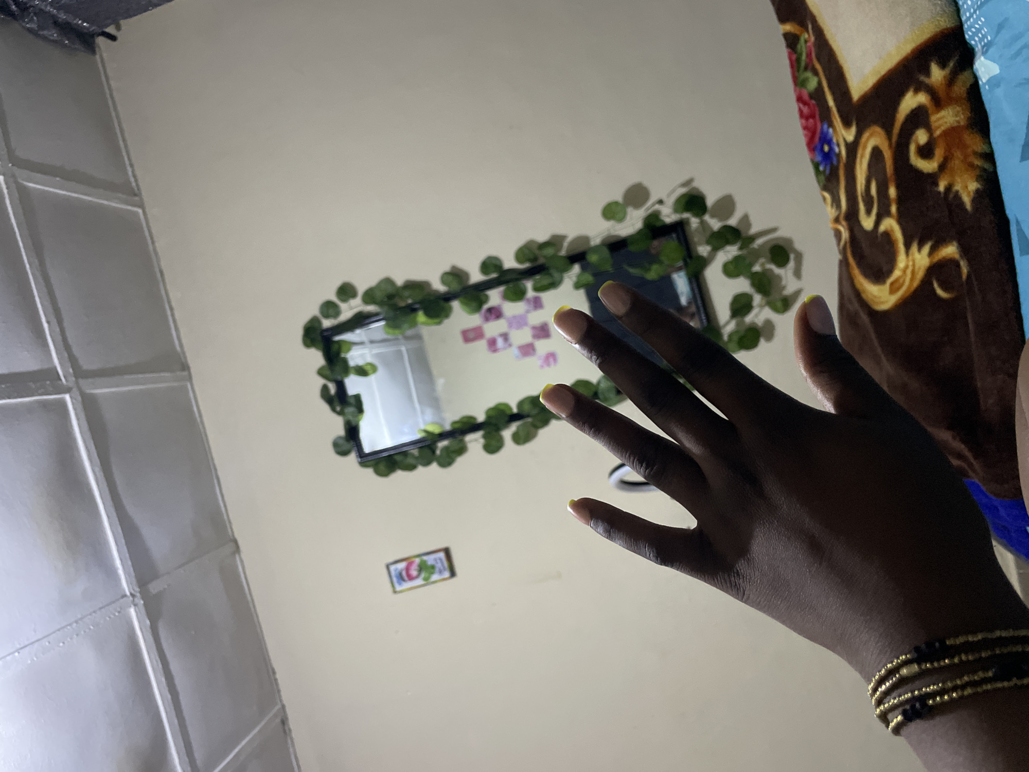 A person's dark-skinned hand with yellow-tipped fingernails and a stack of gold and black beaded bracelets is held up in the foreground, obscuring a portion of a beige wall. A rectangular mirror with a black frame is centered on the wall, decorated with artificial green ivy. Inside the mirror's reflection, a small framed picture is visible on the wall, along with a grid of small pink and white square photographs. The ceiling above is paneled and painted white. In the lower right of the image, a portion of a brown blanket with a gold swirl pattern and floral accents is visible, resting on a blue surface. The overall scene appears to be an indoor domestic setting, likely a bedroom, possibly located in Rafin Par, Nigeria, though specific landmarks are absent.