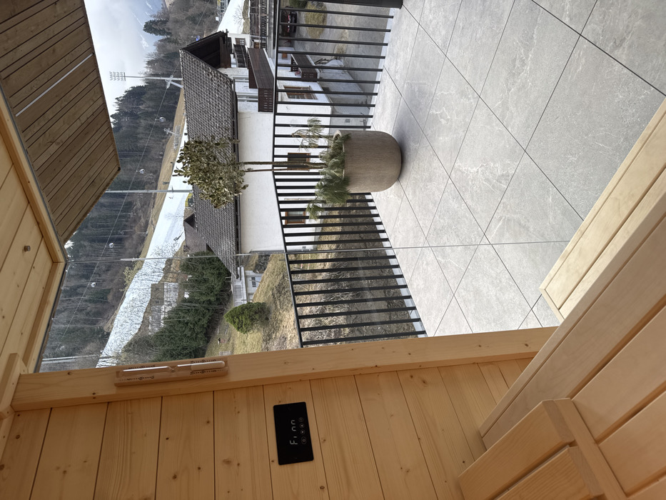 The image displays an interior perspective from a wooden sauna structure, overlooking an outdoor balcony and a mountainous landscape in Schladming, Austria. The immediate foreground shows vertical light-colored wooden planks forming the sauna wall, with two levels of wooden benches extending from the lower left. A black digital control panel, marked "Fionn" with illuminated symbols, is mounted on the wooden wall, positioned below a vertically oriented wooden-cased thermometer and hygrometer.
A transparent glass partition separates the sauna interior from an exterior balcony. The balcony floor is paved with light grey, square tiles featuring a subtle veined pattern. A black metal railing with vertical bars encloses the balcony area. Centered on the balcony is a large, rounded, beige planter containing a small, sparsely foliated tree and some coniferous sprigs at its base. A wooden ceiling overhang extends from the sauna structure above a portion of the balcony.
Beyond the balcony and a grassy embankment, a multi-story white residential building with a brown tiled roof and dark wooden balconies is visible. The background features a prominent, steep mountain slope partially covered with snow and dense evergreen trees. Multiple ski lift gondolas are observed suspended on cables, ascending the snow-covered sections of the mountain. A structure equipped with floodlights, characteristic of a ski resort facility, is present on the upper right side of the mountain. The sky is overcast. No individuals are present in the scene.
