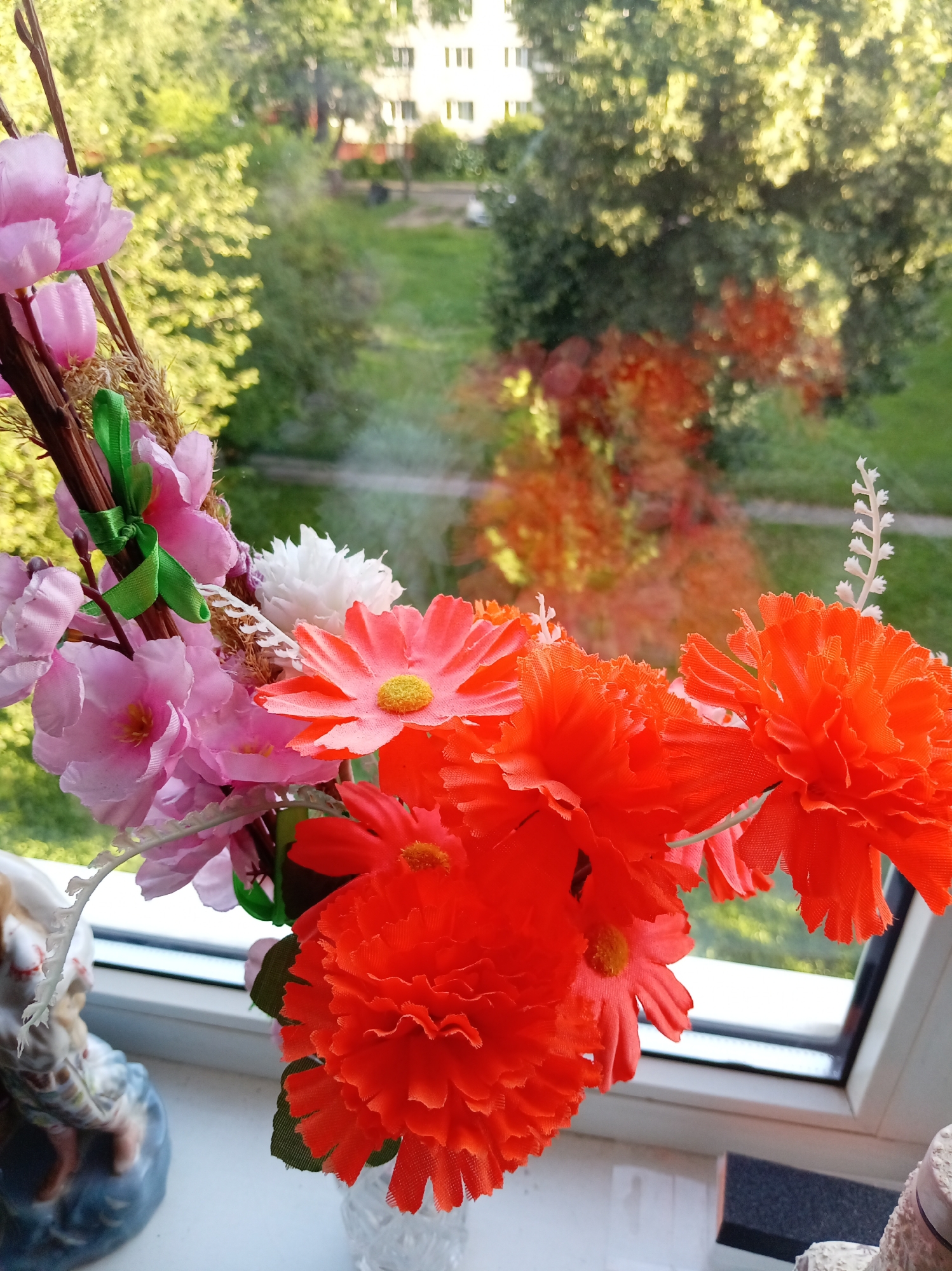 The image shows a bouquet of artificial flowers on a windowsill, with a blurred outdoor scene visible through the window. The flowers are a mix of pink, white, and bright orange. In the foreground, on the left, is a ceramic figurine. The windowsill is white. The outdoor scene depicts green trees, grass, and a building in the background. The lighting suggests it is daytime, likely in the late afternoon due to the warm light. The location is identified as Бараўлянскі сельскі Савет, Belarus.