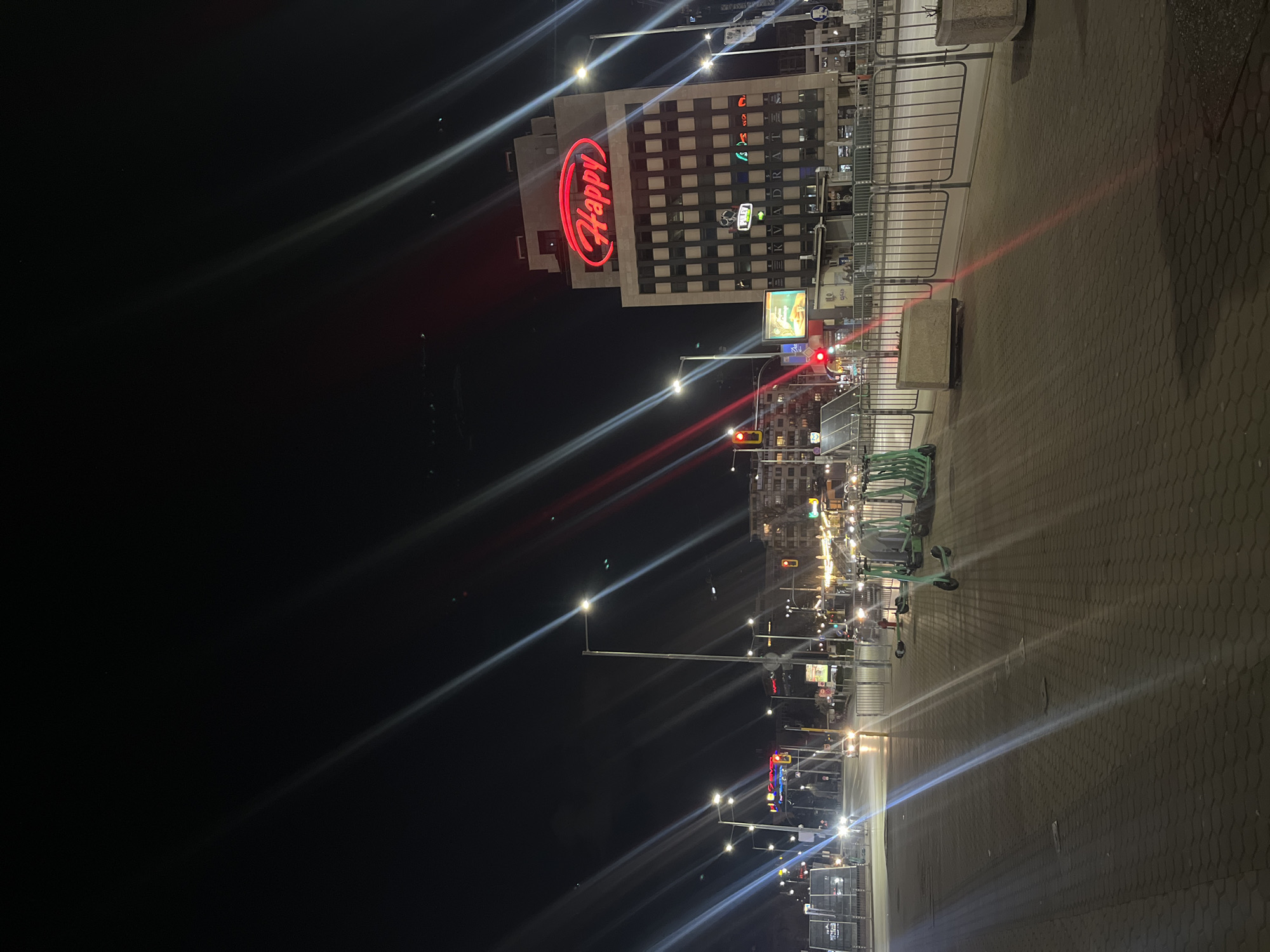 The image depicts a night scene in Sofia, Bulgaria, featuring a street lined with streetlights and several scooters parked on the sidewalk. A building with a large, illuminated "Happy" sign is visible in the background. The sidewalk is paved with interlocking stones, and there are metal barriers along the edge. The street is lit by numerous streetlights, which cast long reflections on the pavement. The sky is dark, and a few other buildings can be seen in the distance, illuminated by their own lights.