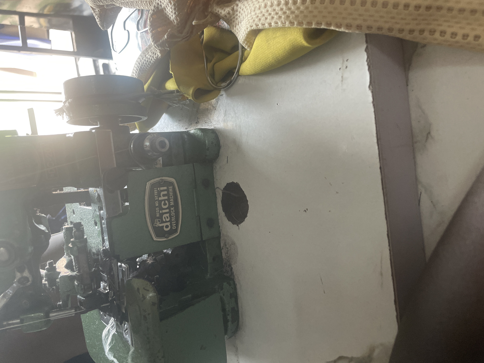 This is a medium shot, eye-level, outdoor photograph taken during the daytime in Jalingo, Nigeria. The image captures a close-up view of a green Daichi overlock sewing machine positioned on a white table, which appears to be worn and stained in places. The machine itself shows signs of use, with some lint and debris visible on its metal components. To the right of the machine, a bright yellow fabric is draped, with a metal ring partially visible. Further to the right, a beige knitted fabric is also present. The background is out of focus, but appears to be an indoor space with bright light coming from an open doorway or window, suggesting a workshop or domestic setting. The overall impression is one of a functional, perhaps well-used, sewing station.
