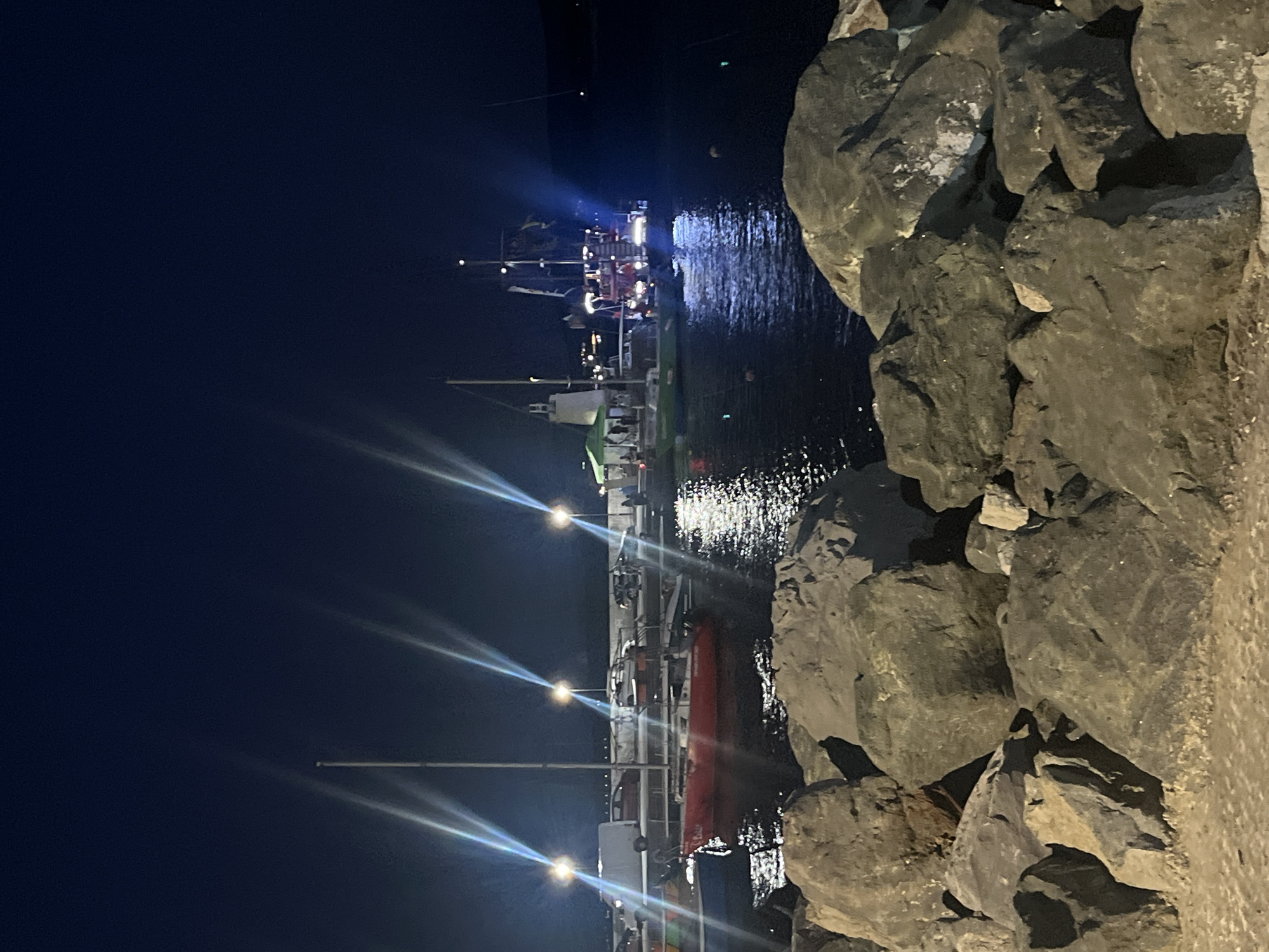 This image captures a nighttime scene at a marina in Primorsko, Bulgaria. In the foreground, a breakwater constructed of large, dark rocks is visible, with a sandy or gravelly surface in the very bottom foreground. Beyond the rocks, a body of dark water reflects the lights from the shore and from vessels docked in the marina.
Several boats are present. A red sailboat with a white cabin is docked in the middle ground, its mast rising vertically. To its left, another boat, partially obscured, appears to be a dark blue color. Further into the marina, a larger, more elaborate boat resembling a pirate ship with multiple masts and sails is illuminated. Its decks are lit, and it appears to be a passenger vessel. A green canopy is visible on a smaller boat to the left of the pirate ship, with several people standing underneath it.
Along the shoreline in the background, a promenade or boardwalk is visible, with several vehicles parked, including a white van and other cars. Streetlights illuminate the area, casting distinct beams of light into the night sky. The overall atmosphere is one of a lively evening at a coastal town's harbor.