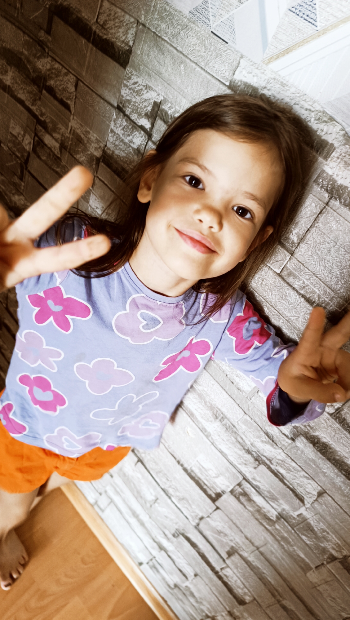 A young girl with long, dark hair is lying down and looking up at the camera. She is smiling and holding up two peace signs with her hands. She is wearing a light blue long-sleeved shirt with a floral pattern of pink and purple flowers, and bright orange shorts. The background appears to be a brick-patterned wallpaper, and to the lower left, there is a wooden floor. The lighting suggests it is daytime, possibly indoors. The girl's expression is happy and playful. No text is visible in the image. The scene is set in Karsha, Russia.