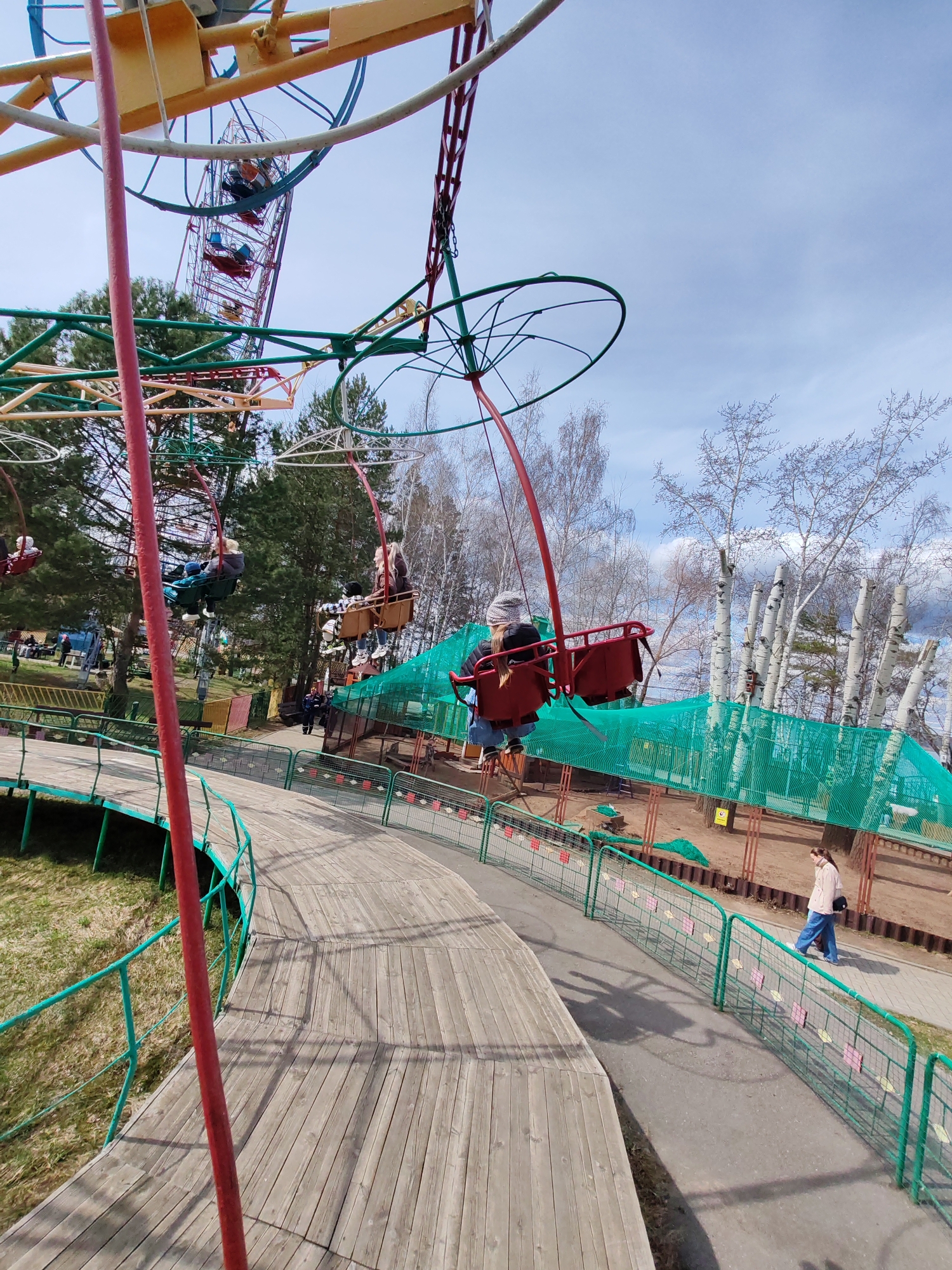 The image depicts an outdoor scene in Chaikovsky, Russia, featuring amusement park rides and elements. The main focus is a swing ride with red seats, a green frame, and occupants, moving in a circular motion. In the background, a Ferris wheel is visible. A wooden walkway with a green railing curves through the scene, and a person is walking along a paved path parallel to the walkway. The environment includes trees with bare branches and green netting enclosing a sandy area. The sky is partially cloudy.