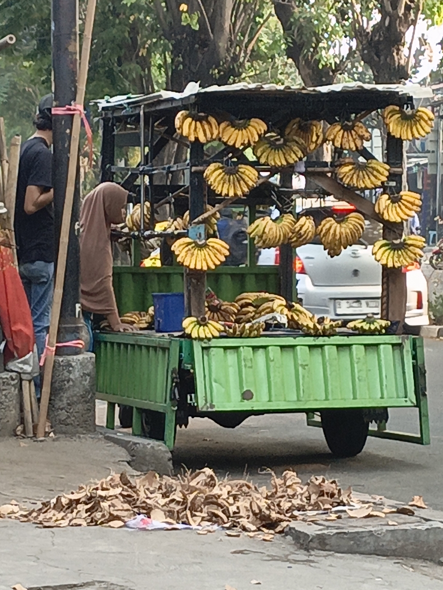 A street vendor in East Jakarta, Indonesia, is seen selling bananas from a green cart. The cart is loaded with bunches of yellow bananas, some of which are displayed hanging from the roof. The vendor, a woman wearing a brown hijab, is standing behind the cart, possibly interacting with a customer who is partially visible on the left side of the image. The background shows trees, a white car, and more foliage, suggesting a street scene in a tropical urban environment. A pile of dry leaves is visible on the pavement in the foreground. The scene appears to be set on a paved street next to a sidewalk.