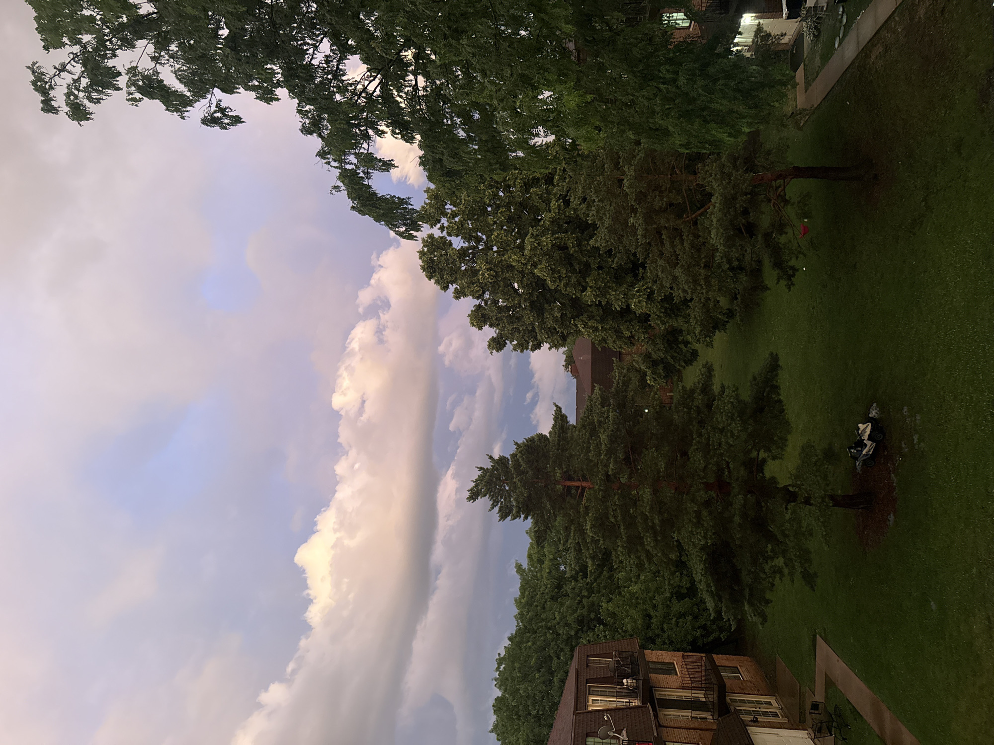 The image captures a dusky sky over an urban residential area. In the foreground, two large pine trees stand on a grassy lawn, with a white all-terrain vehicle parked near the base of the right tree. Adjacent to the lawn, a brick apartment building with brown roofs and balconies is visible on the left, and another similar building is partially seen on the right. Dense green foliage surrounds the buildings and the courtyard. The sky is filled with dramatic clouds, illuminated by the fading light of dusk, with hints of pink and purple hues. The scene appears to be a quiet evening in a suburban setting, likely in Glendale Heights, USA.