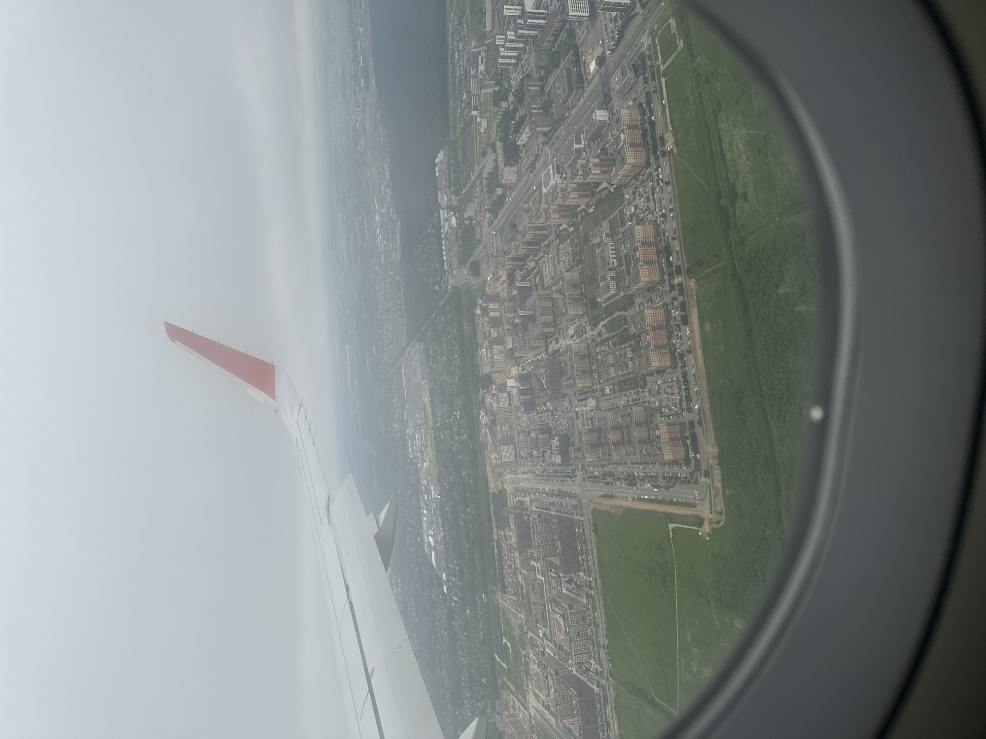 This is an aerial photograph taken from an airplane window, looking down on the city of Slavyanka, Russia. The image is characterized by a hazy, overcast sky, which contributes to a muted color palette. The aircraft's wing, painted white with a red tip, occupies the upper left portion of the frame, extending diagonally across the sky.
Below the wing, a dense urban landscape unfolds. Numerous apartment buildings, characterized by their reddish-brown facades and rectangular shapes, are visible. These buildings are clustered together, forming distinct neighborhoods. Streets and roads weave through the cityscape, with cars parked along them, indicating a populated area.
Adjacent to the built-up areas are large expanses of green, likely parks, fields, or undeveloped land. These green spaces provide a contrast to the man-made structures. In the distance, more buildings and greenery can be seen, fading into the hazy atmosphere. The overall impression is of a modern, developing city under a gray, cloudy sky. The view is framed by the circular airplane window, which occupies the bottom portion of the image.