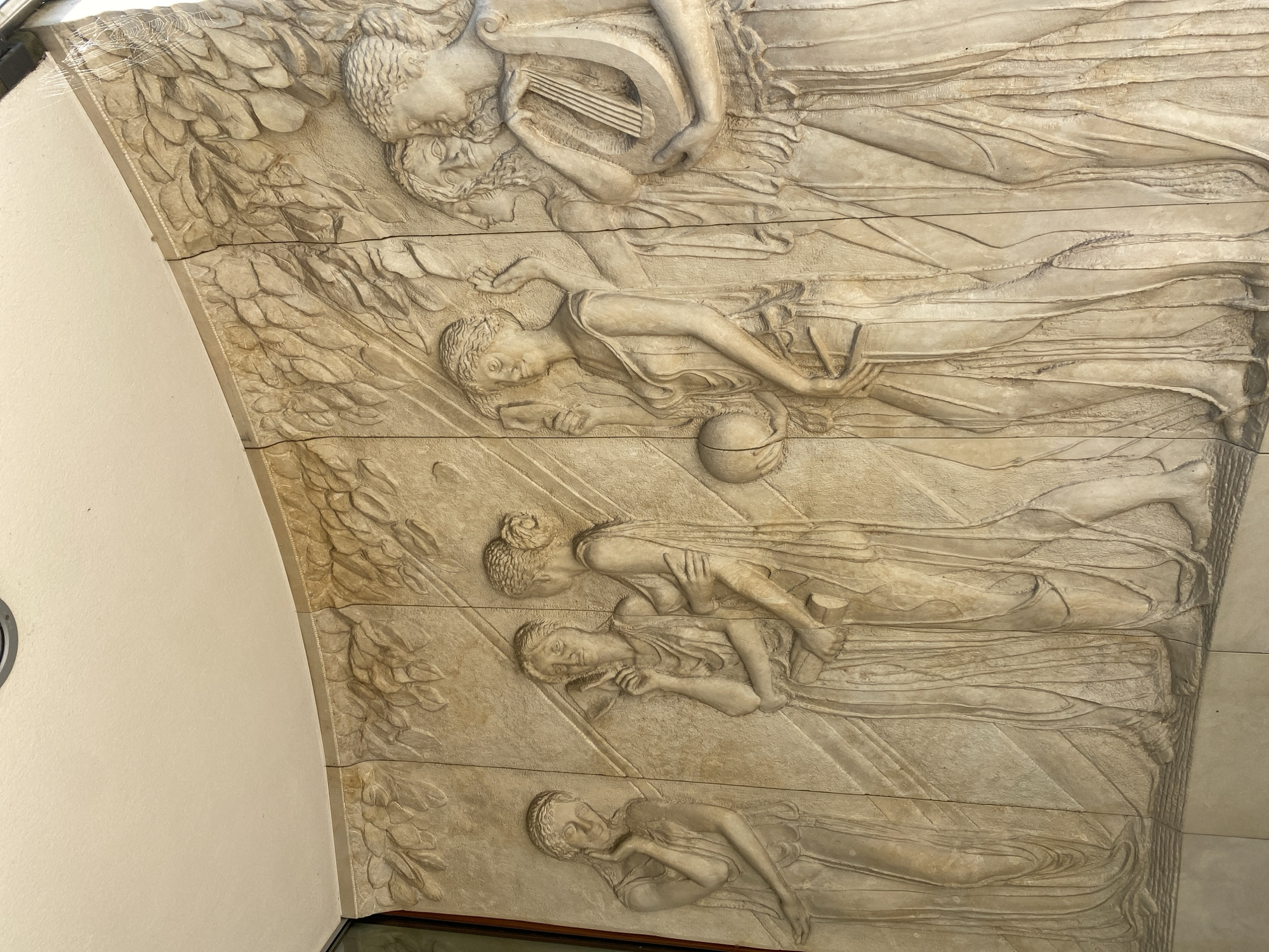 The image shows a bas-relief carving on a curved wall. The carving depicts several female figures in classical attire, arranged in a frieze-like composition. The figures are rendered in a stylized manner, with flowing drapery and idealized features. The background is textured with a pattern resembling leaves and branches. The relief is made of a light-colored stone or plaster and is mounted on a wall, likely an exterior facade, given the presence of what appears to be an overhang or ceiling above it. The carving is detailed, with individual strands of hair and facial features clearly defined. The figures are interacting with each other, some are looking at each other, while others appear to be engaged in individual activities. One figure on the right is playing a lyre. The overall impression is of a classical or neoclassical artwork. This specific bas-relief is located on the facade of the National Museum in Warsaw, Poland.