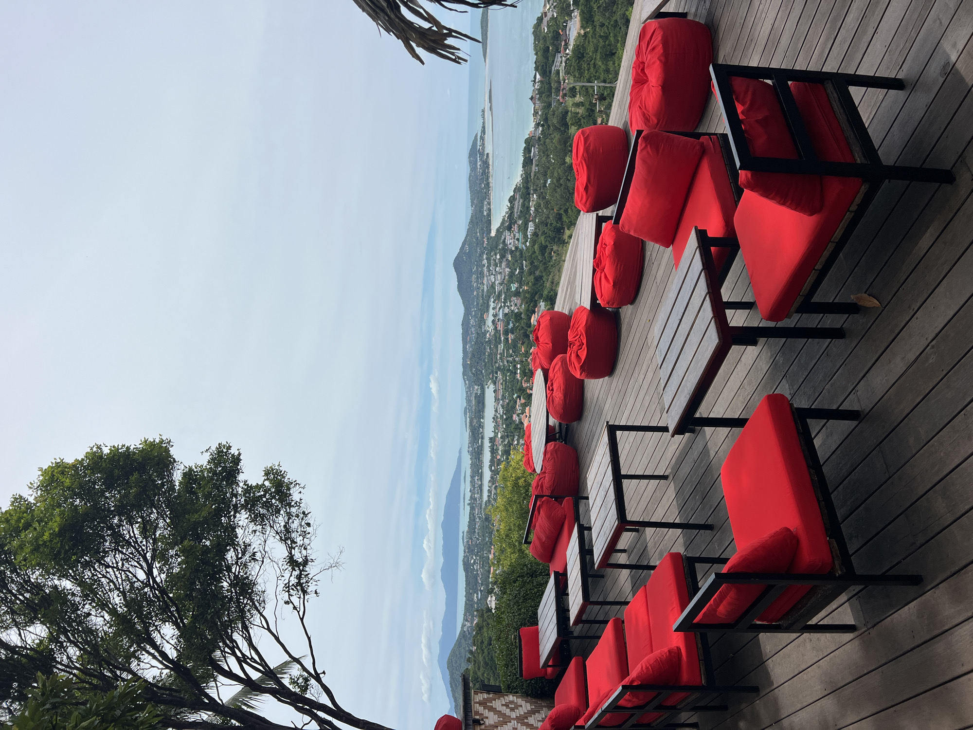 This image captures an outdoor seating area on a wooden deck, offering a panoramic view of Baan Chaweng Noi, Thailand. The seating consists of several red chairs with black frames and red cushions, interspersed with wooden tables. Additionally, several large red beanbag chairs are scattered across the deck, providing casual seating options. In the background, a lush green landscape descends towards the sea, with the town of Baan Chaweng Noi visible along the coastline. The horizon is dominated by mountains and a pale, overcast sky. A large tree with dense foliage is present in the upper left corner of the frame, and a portion of a thatched roof is visible in the upper right. The scene appears to be set during the daytime, with diffused light. There are no people visible in the image.