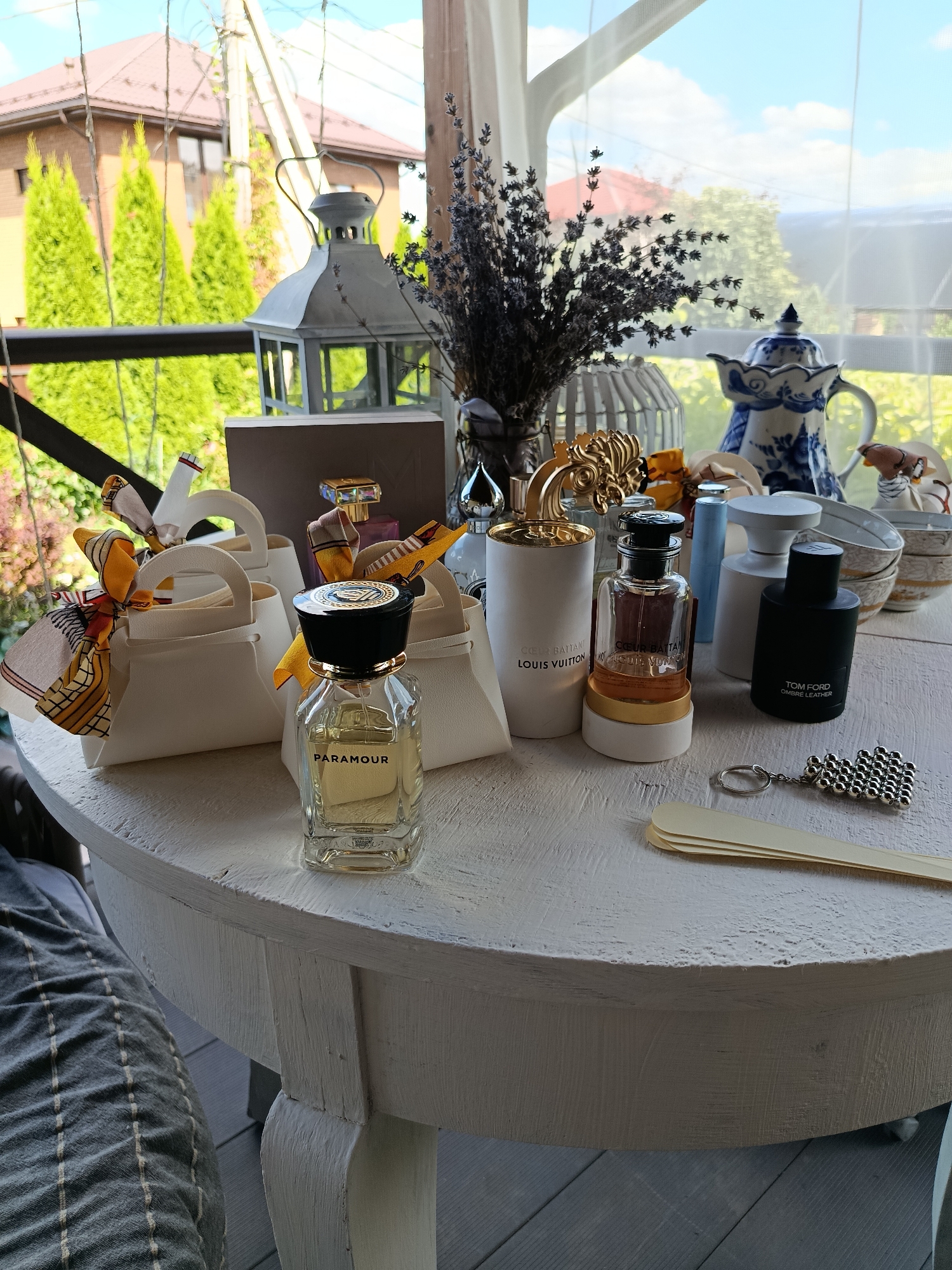 The image depicts an outdoor setting, likely a patio or balcony, with a white, round table as the central focus. On the table, there is a collection of perfume bottles, decorative items, and small white gift bags. In the foreground, a bottle labeled "PARAMOUR" is prominent. Behind it, several white gift bags adorned with colorful scarves are arranged. To the right, there are other perfume bottles, including one labeled "LOUIS VUITTON" and another labeled "TOM FORD OMBRÉ LEATHER." Various decorative objects are scattered around, including a blue and white ceramic teapot, a lantern, dried lavender, and what appears to be a decorative gilded birdcage. A keychain with a metallic sphere charm and some wooden spatulas are also on the table. In the background, out-of-focus trees, houses, and a telephone pole are visible, suggesting a residential area. The overall ambiance is one of organized display, possibly for a gift presentation or a product showcase. The location is identified as Мосметаллоконструкция Онт, Russia, indicating an urban or suburban environment. There are no people visible in the image.