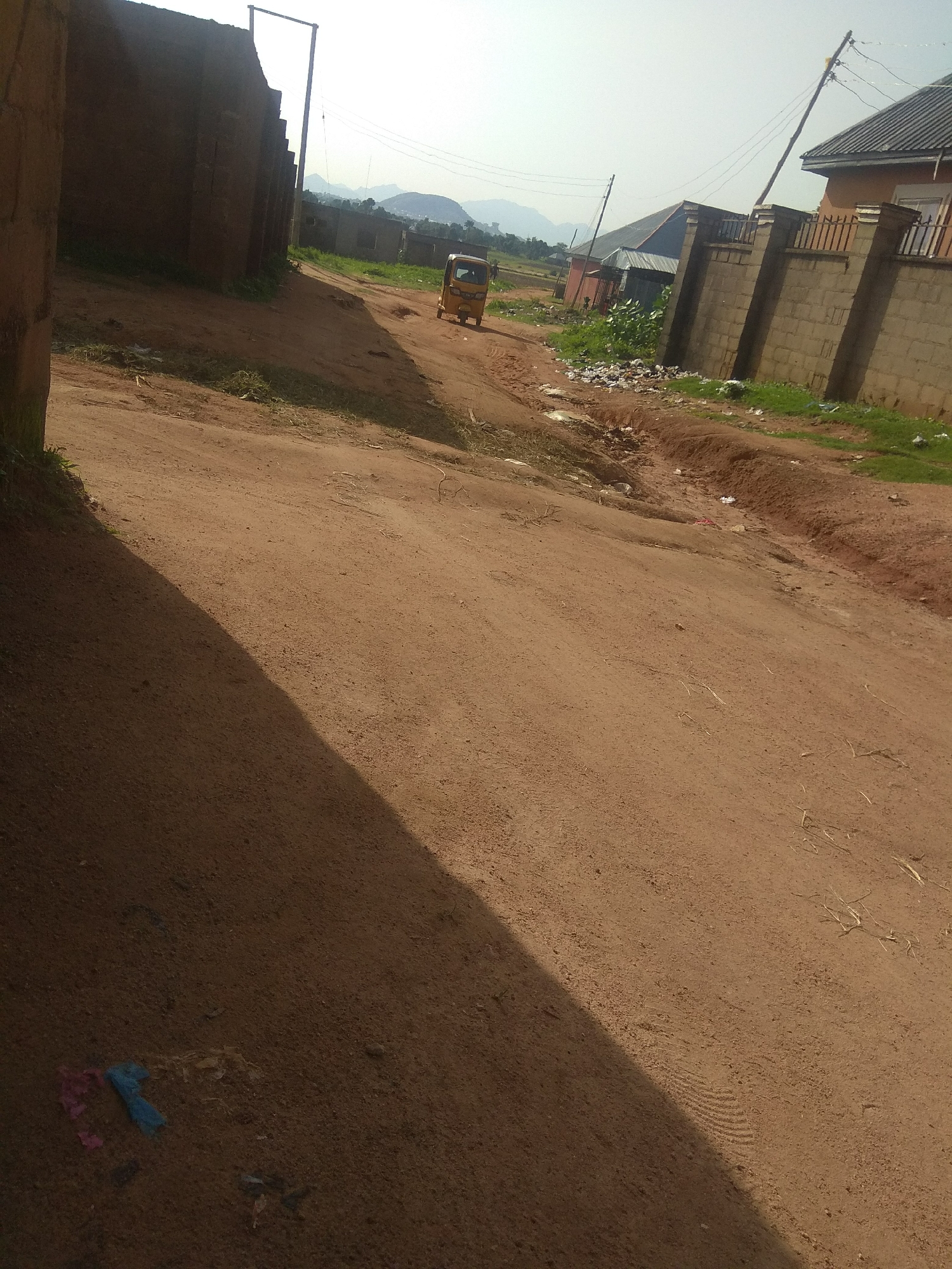 A dirt road in a residential area of Jalingo, Nigeria. A yellow auto-rickshaw is visible on the road. To the left of the road is a brick wall and the side of a building. To the right, there are more buildings with fenced yards and a power pole. The ground is mostly dirt with some grass and debris. The sky is overcast.
