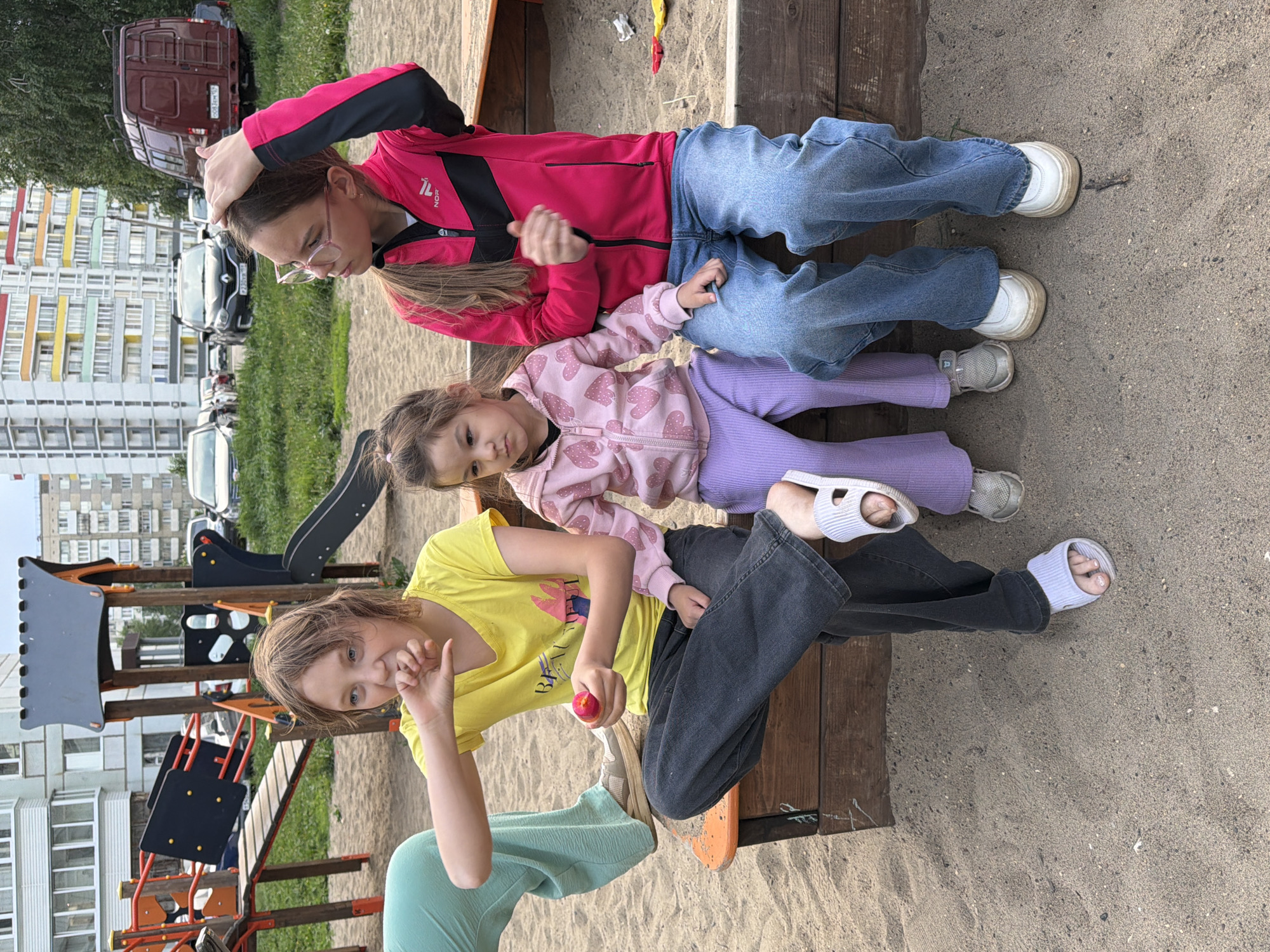 Three young girls are sitting on a wooden bench in what appears to be a children's playground in Chaikovsky, Russia. The playground is located near apartment buildings.
The girl at the top, wearing glasses, has a pink and black jacket and blue jeans. She is touching her hair. The middle girl has a light pink jacket with hearts, purple pants, and white sandals. The girl at the bottom wears a yellow shirt, dark pants, and white sandals. She is holding a small red object, possibly an apple or a toy, in her hand, and is looking at the camera. The other half of the girl is wearing green pants.
The playground includes a slide and other climbing structures made of wood and metal. The ground is covered in sand. Vehicles and apartment buildings are visible in the background.