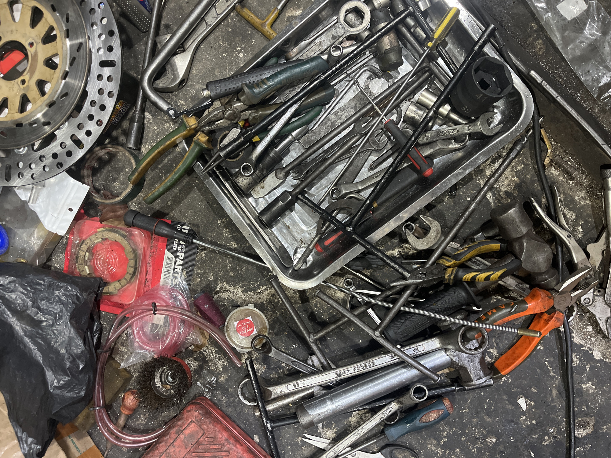 An overhead, high-angle shot captures a chaotic workbench cluttered with a variety of tools and motorcycle parts. The scene is set in a workshop located in Tangerang, Indonesia. Various hand tools, including wrenches, pliers, screwdrivers, and a socket set, are scattered across the dirty, concrete surface. A large, perforated brake rotor is positioned in the upper right quadrant of the image. Several red plastic containers and a black plastic bag are also visible, adding to the disarray. A small, circular container with a red and white sticker labeled "Hi-Temp CV Grease" is present among the tools. The lighting is dim, casting shadows and highlighting the metallic sheen of the tools. No people are present in the image.