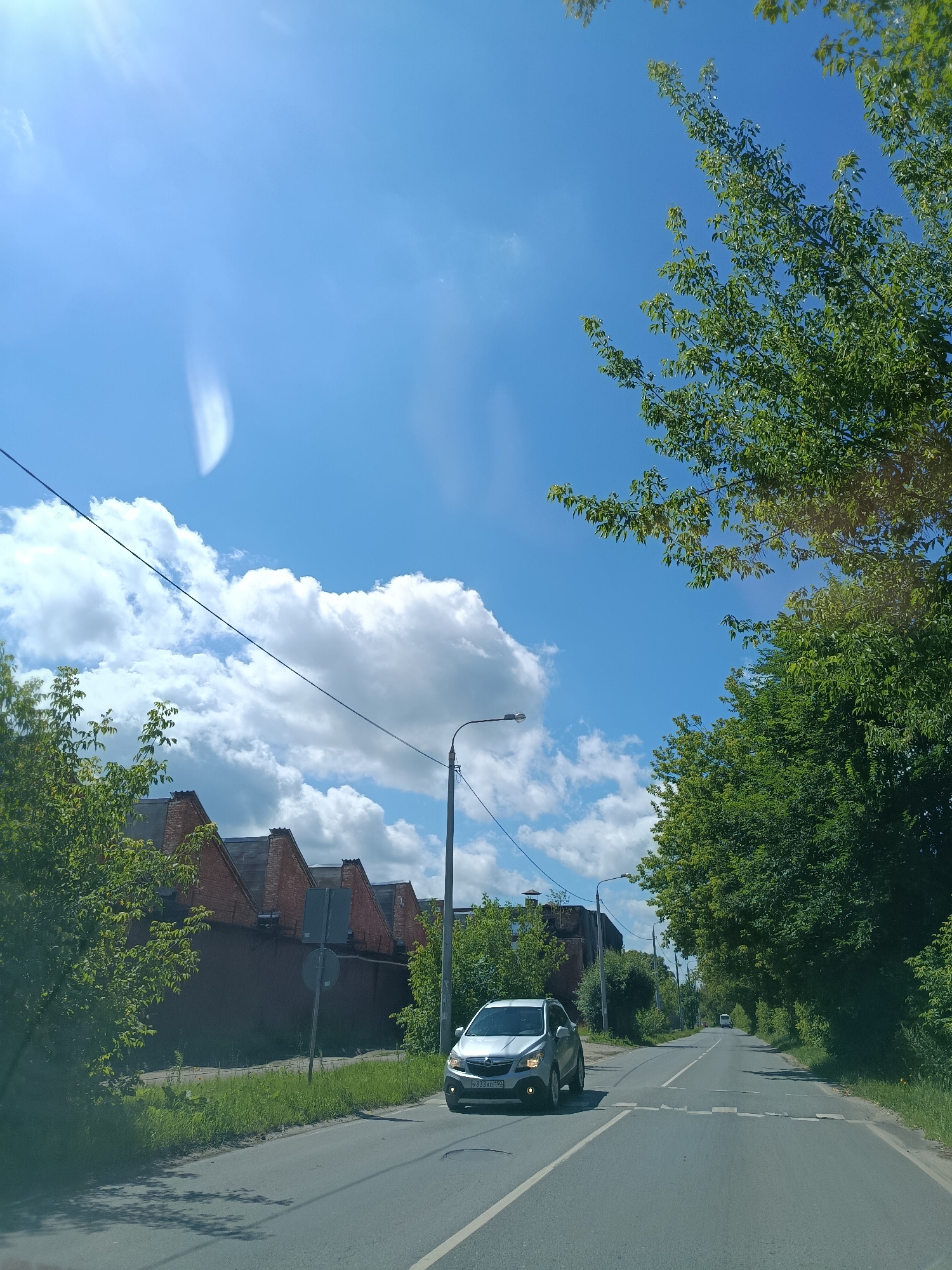 The image depicts a street scene in Orekhovo-Zuyevo, Russia, on a bright, sunny day with scattered clouds. The perspective is from inside a vehicle, looking forward along a paved road.
On the left side of the road, there is a long, low brick building with a dark, corrugated metal facade and a series of red brick gables. This building is partially obscured by lush green trees and foliage. A grey traffic sign is visible in front of the building.
In the foreground on the left, a silver hatchback car, possibly a Opel Mokka, is driving on the road. Its license plate is visible and appears to read "У333ХО 150".
The road itself is a two-lane asphalt surface with white lane markings. There are pedestrian crossing markings painted in white dashes across the road ahead of the silver car.
On the right side of the road, dense green trees and foliage line the way, creating a verdant border. A lamppost is visible along the roadside, and another one is further down the road. In the distance, a white van or bus can be seen on the road.
The sky is a vibrant blue, with large, puffy white clouds. Sunlight streams in from the upper left, creating lens flare effects in the image. The overall impression is one of a suburban or semi-rural setting on a pleasant summer day.