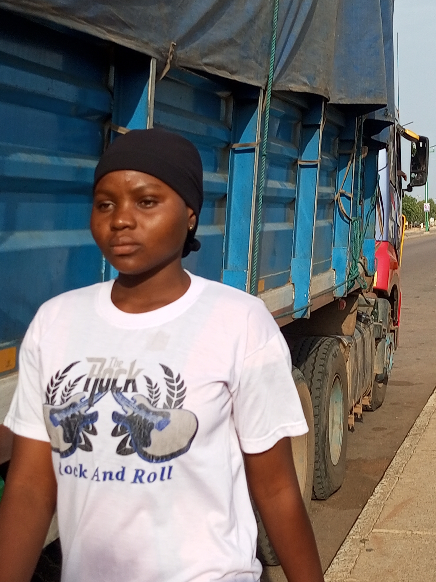 A young woman with dark skin and dark hair, wearing a black headscarf and a white t-shirt with a "Rock and Roll" graphic featuring two bulls, stands in front of a large blue truck. The truck has a covered bed and is parked on a paved surface next to a sidewalk. The woman is looking slightly to her right, and the truck's tires are visible on the right side of the image. The scene appears to be outdoors, with a clear sky in the background. The image was taken in Wuro Dole, Nigeria.