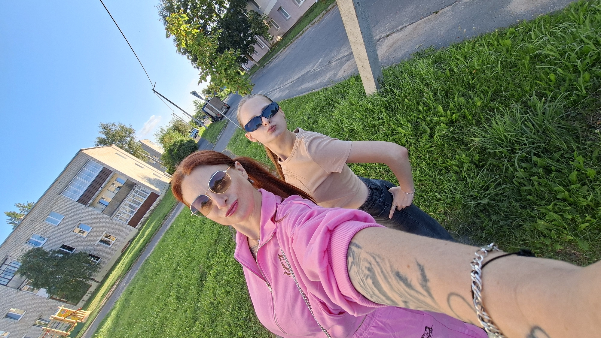 Two women are posing for a selfie in a grassy area next to a paved road. The woman in the foreground is wearing a pink tracksuit and sunglasses. She has red hair styled in a ponytail and a bracelet on her wrist. The woman behind her is wearing a light-colored t-shirt and dark jeans, also wearing sunglasses. She has her hair in a ponytail as well. They are standing in front of a light-colored apartment building. The sky is clear and blue. The scene appears to be in the city of Myadzyel, Belarus, with typical apartment buildings and green spaces found in Eastern European cities.