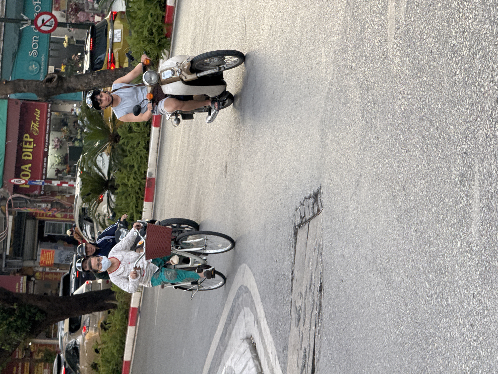 In Hà Nội, Vietnam, on a street with a curved asphalt road, two individuals are traveling on vehicles. On the left, a woman rides a bicycle with a basket on the front, wearing a mask and light-colored patterned shirt with teal pants. Behind her, a person on a motorcycle wears a helmet. On the right, a young man on a beige and brown scooter wears goggles, a gray sleeveless shirt, and shorts. Traffic signs and buildings are visible in the background.