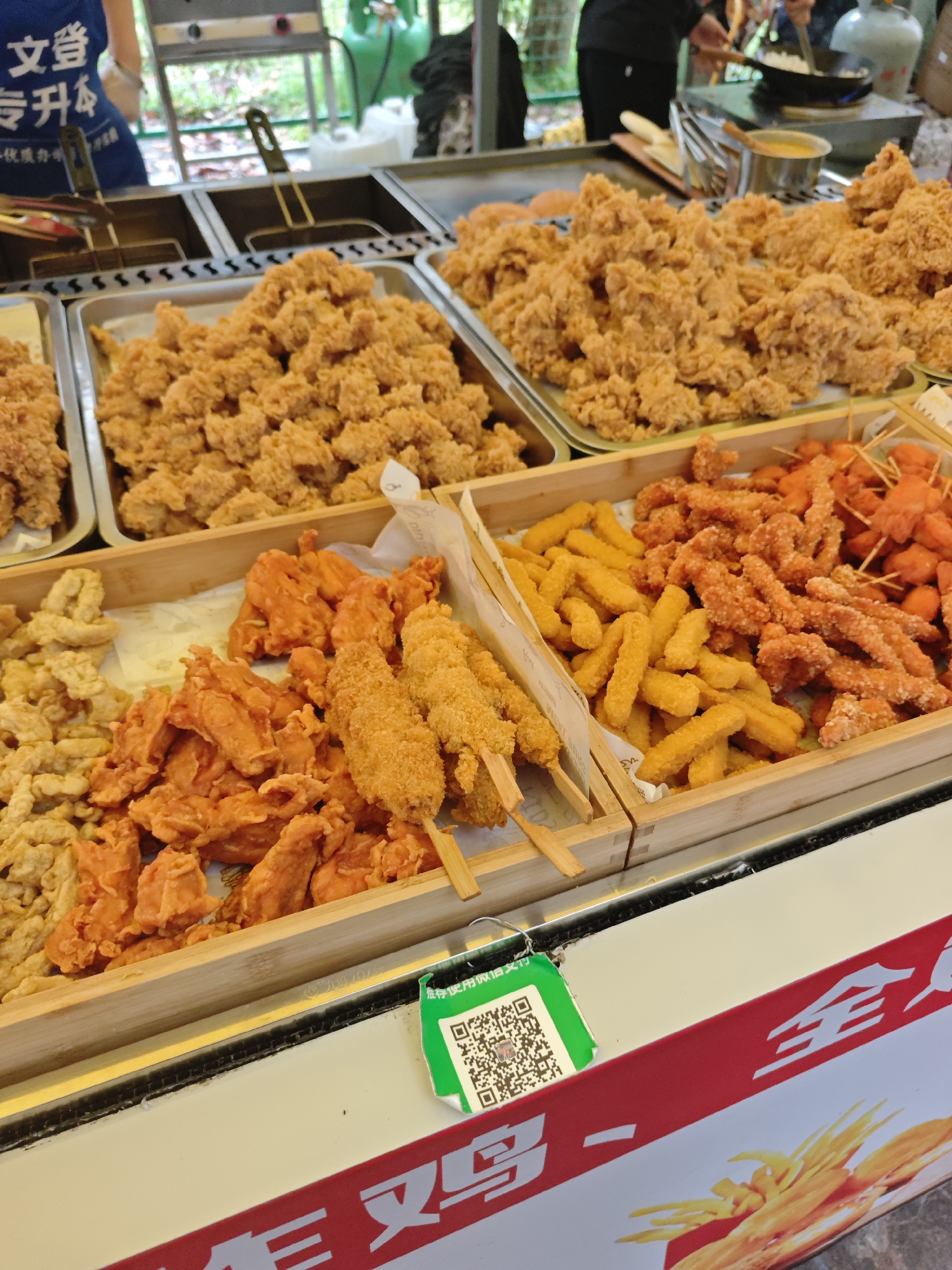 A vendor stall in Shapingba District, China, displays a variety of fried foods in metal trays and wooden containers. The trays contain piles of golden-brown, irregularly shaped fried pieces. The wooden containers hold an assortment of items, including light-colored fried bits, orange-colored fried chunks, and skewers with breaded and fried foods. A container also contains yellow fried sticks and orange fried pieces on sticks. A green and white QR code tag hangs from the front of the stall, which has a red and white sign with food imagery and Chinese characters. People are visible in the background.