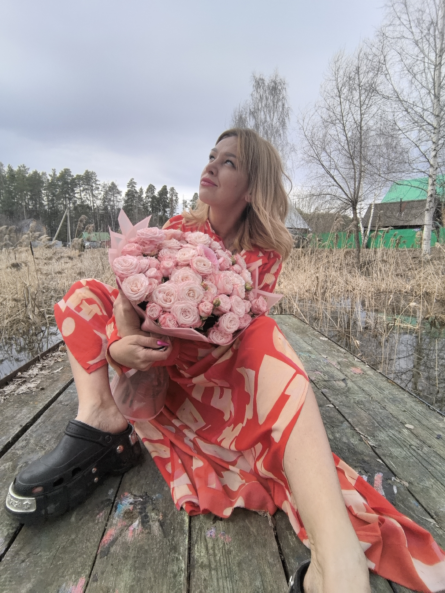 A full-length shot features a blonde woman sitting on a wooden dock, holding a bouquet of light pink roses. She is wearing a long, red and cream patterned dress and a black platform clog shoe. Her gaze is directed upward and slightly to the left.
The dock is situated near a body of water, reflecting the surrounding vegetation and sky. Behind the woman, there is a marshy area with tall, dry reeds. In the background, there are trees, and a building with a green roof is visible. The sky is overcast.
The image was likely taken in a rural area, possibly in the city of Noviy, Russia.