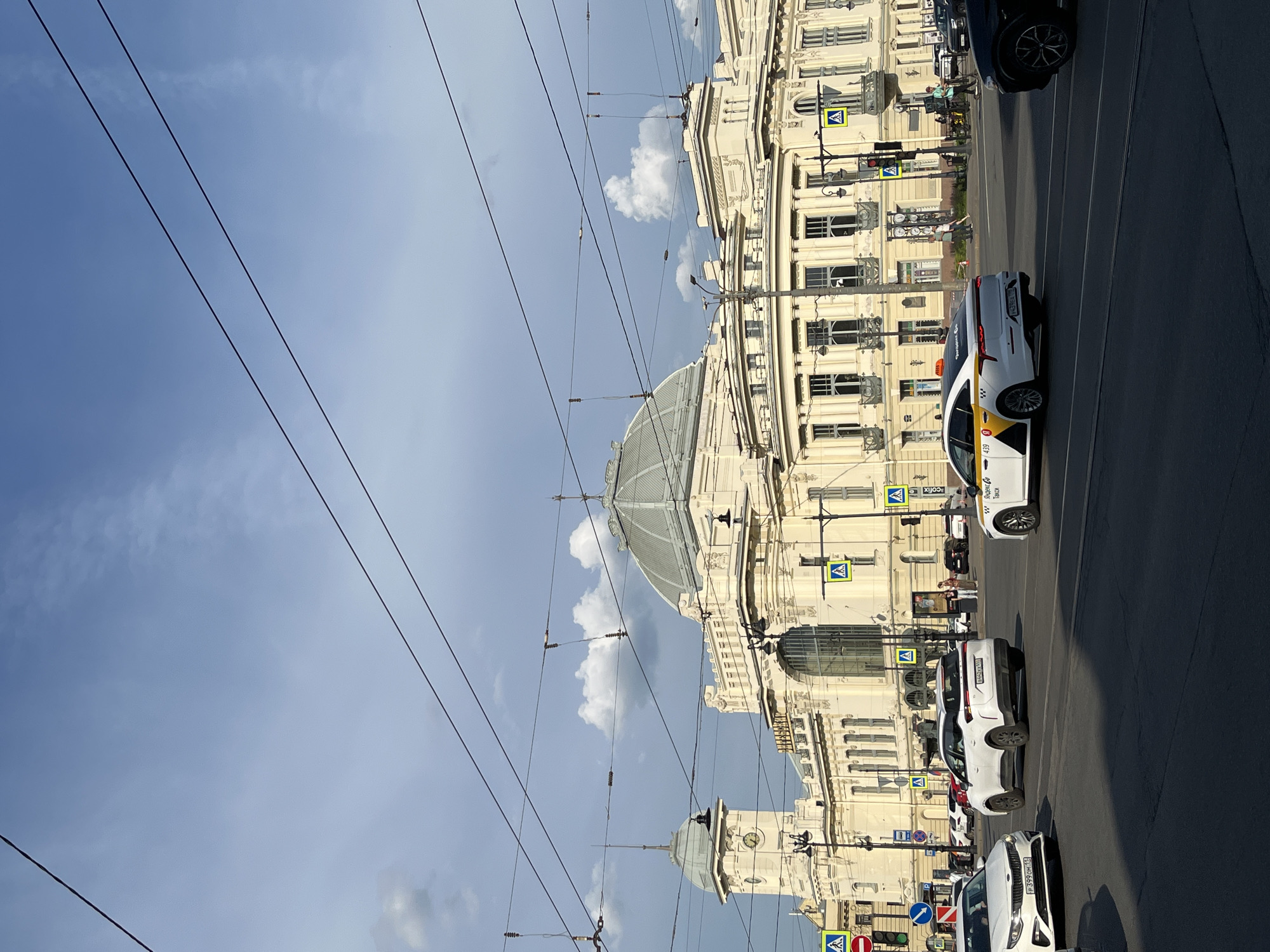 This detailed image captures Moskovsky railway station in Saint Petersburg, Russia, under a partly cloudy sky. The grand, light-colored building, characterized by its prominent central glass dome and a clock tower on the left, dominates the background. The facade features classical architectural elements, including arched windows, decorative cornices, and pilasters.
Numerous black overhead electric wires, likely for trams or trolleybuses, crisscross the blue sky, which contains scattered white cumulus clouds. Some clouds appear darker, suggesting varying weather conditions.
In the foreground, a multi-lane asphalt road occupies the lower portion of the image. Several vehicles are in motion:
* On the far left, a white Kia K5 sedan with Russian license plate "P399OH 178" is partially visible.
* In the center, a white SUV with black wheel arches and Russian license plate "H442KA 198" is moving away from the viewer.
* To its right, a white Kia K5 sedan, operating as a "Yandex Go Taxi" with a yellow and black livery and Russian license plate "P439EA 198," is also heading away.
* A dark-colored SUV is partially visible on the far right.
Traffic infrastructure includes multiple pedestrian crossing signs (blue squares with white triangles), a red "no entry" sign, and a blue circular sign indicating a mandatory right turn. Several lampposts are positioned along the street and sidewalks. Pedestrians are visible on the sidewalks and crosswalks in the mid-ground, though their individual details are indistinct due to distance. The scene depicts a bustling urban environment typical of a major city center.
