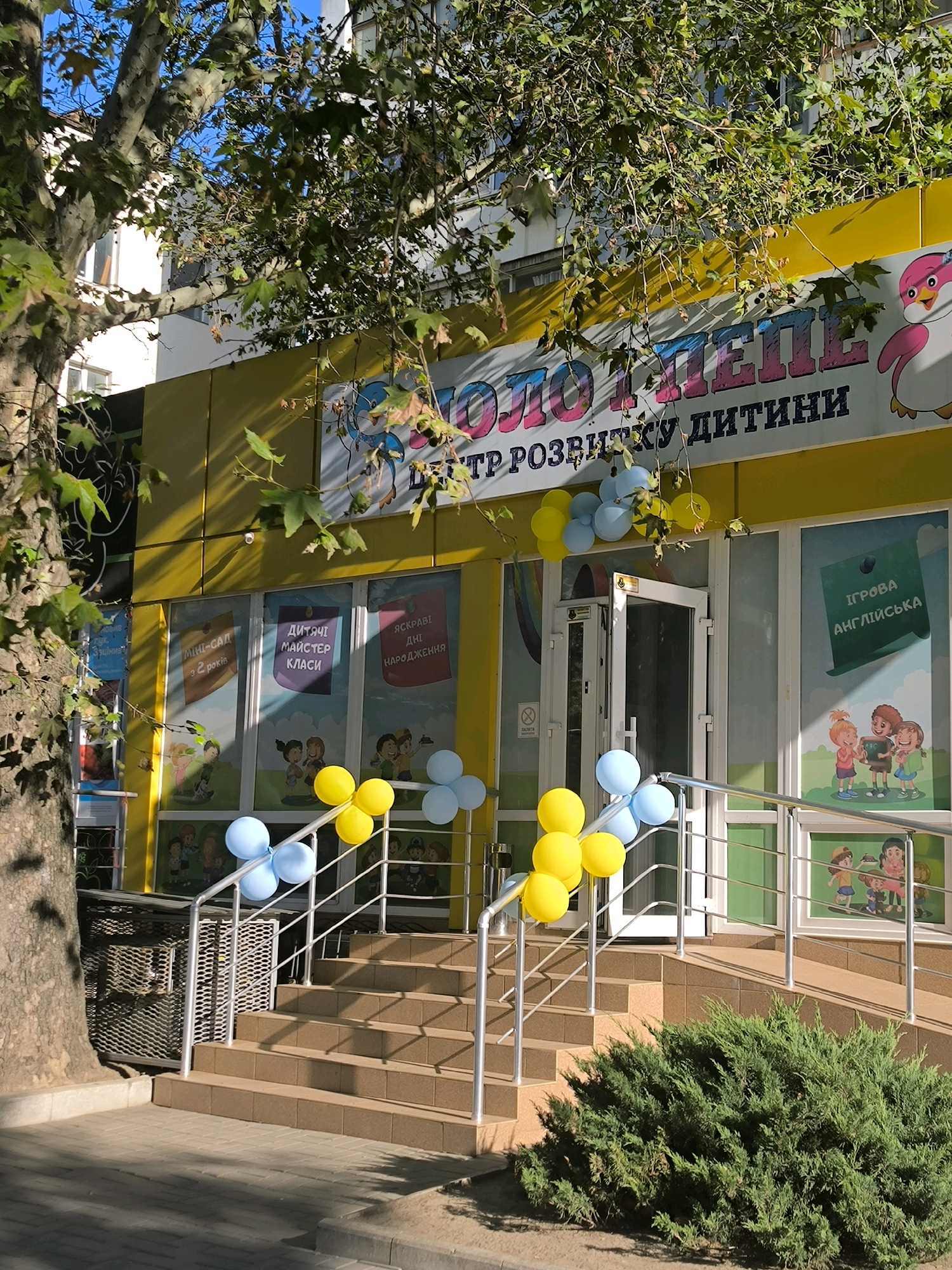 The image shows the exterior of a children's development center called "ЗОЛОТИЙ КЛЮЧИК" in Mykolaiv, Ukraine. The building is painted yellow and features a large sign with the center's name in colorful lettering and a cartoon penguin. The entrance is a glass door flanked by windows displaying children's activities. A set of stairs leads up to the entrance, decorated with blue and yellow balloons. A large tree with green leaves partially obscures the building. The scene appears to be in daylight, with sunlight casting shadows on the ground.